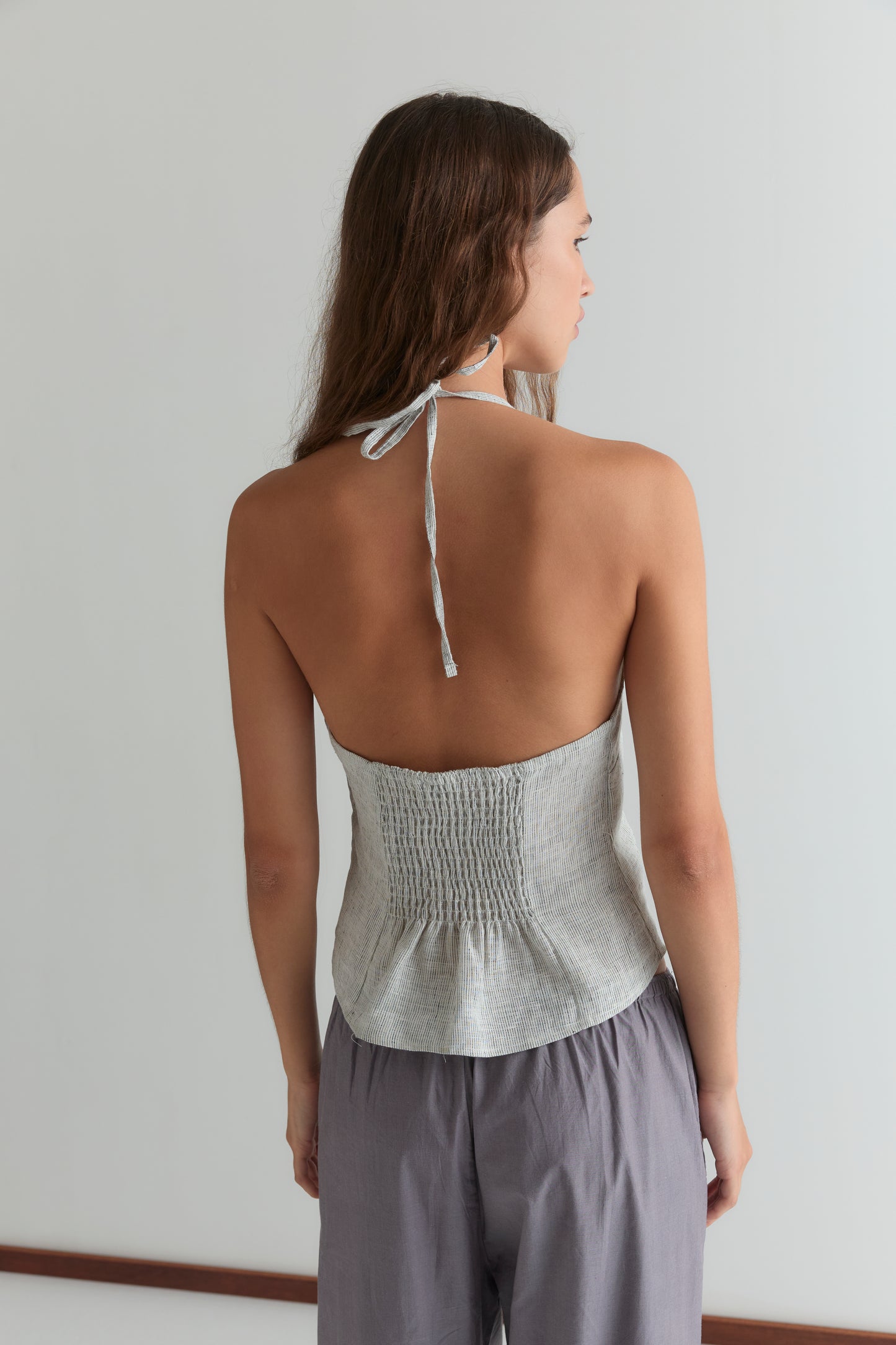 A woman with long brown hair stands with her back to the camera, wearing Deiji Studios' Close Halter Top - Fine Stripe and loose, light gray pants in a minimal indoor setting.