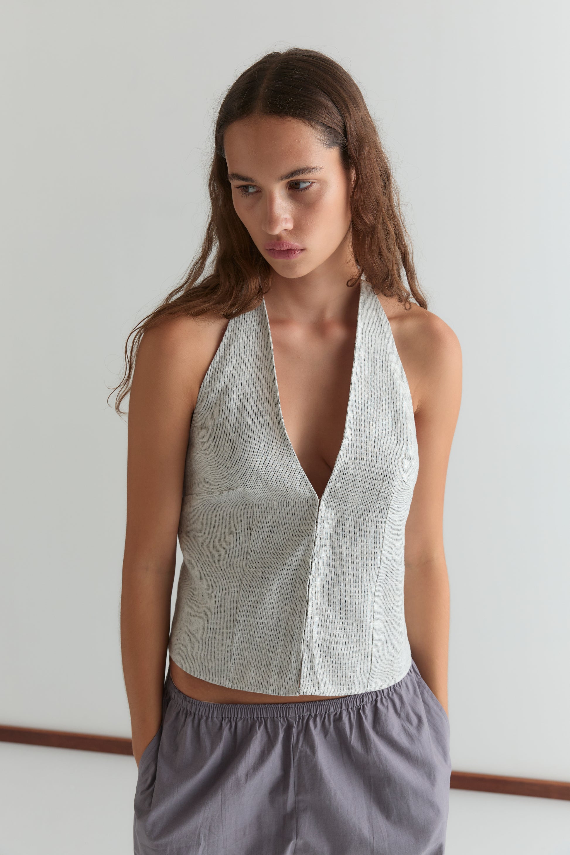 A woman with long brown hair wears Deiji Studios' Close Halter Top - Fine Stripe and loose striped gray pants, standing against a plain white background with her hands in her pockets.