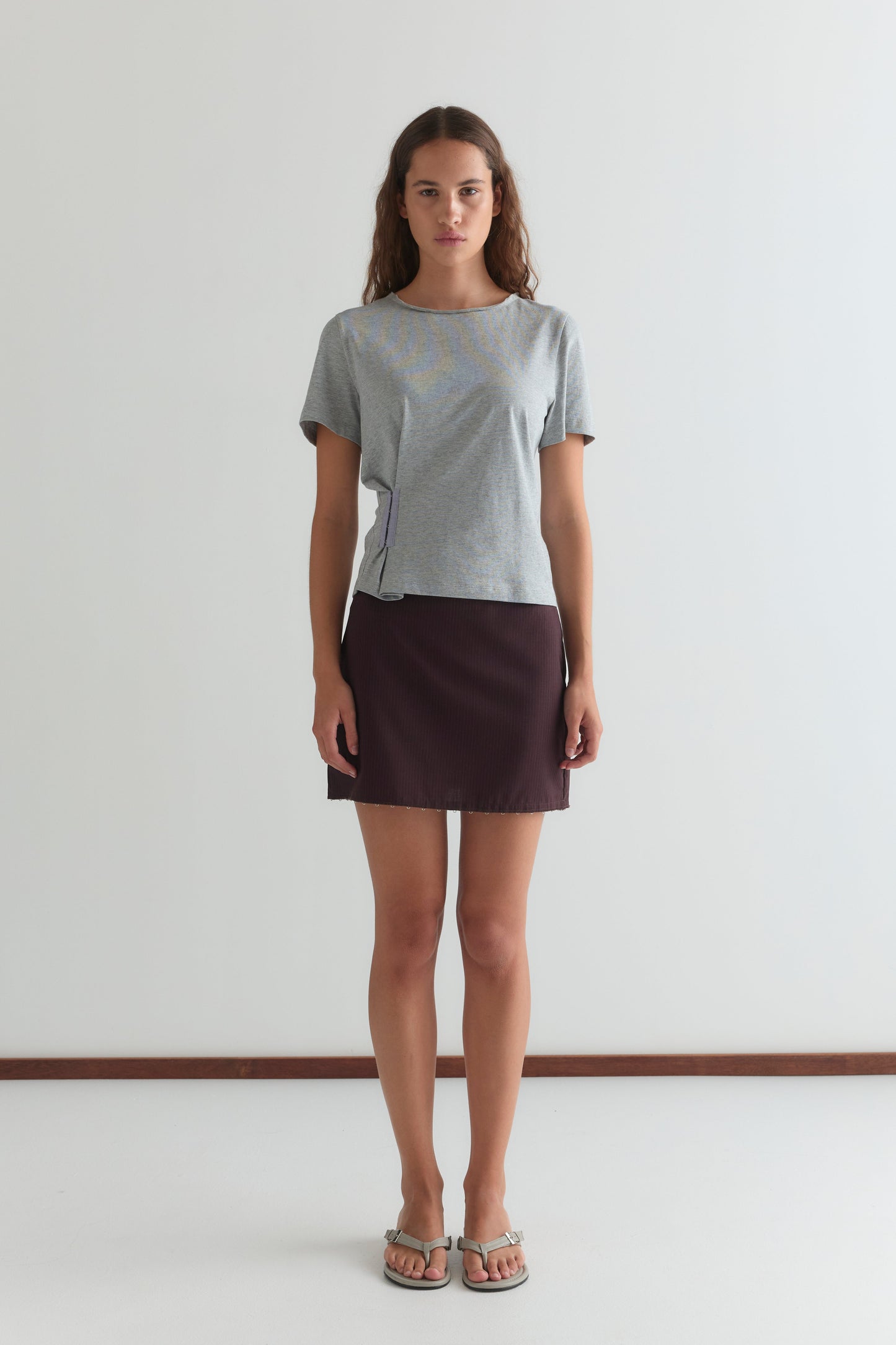 A woman with long, wavy brown hair and a neutral expression stands facing forward against a plain white wall, wearing a light gray short-sleeve t-shirt, the Eye Skirt - Burgundy Brown Stripe, and sandals.