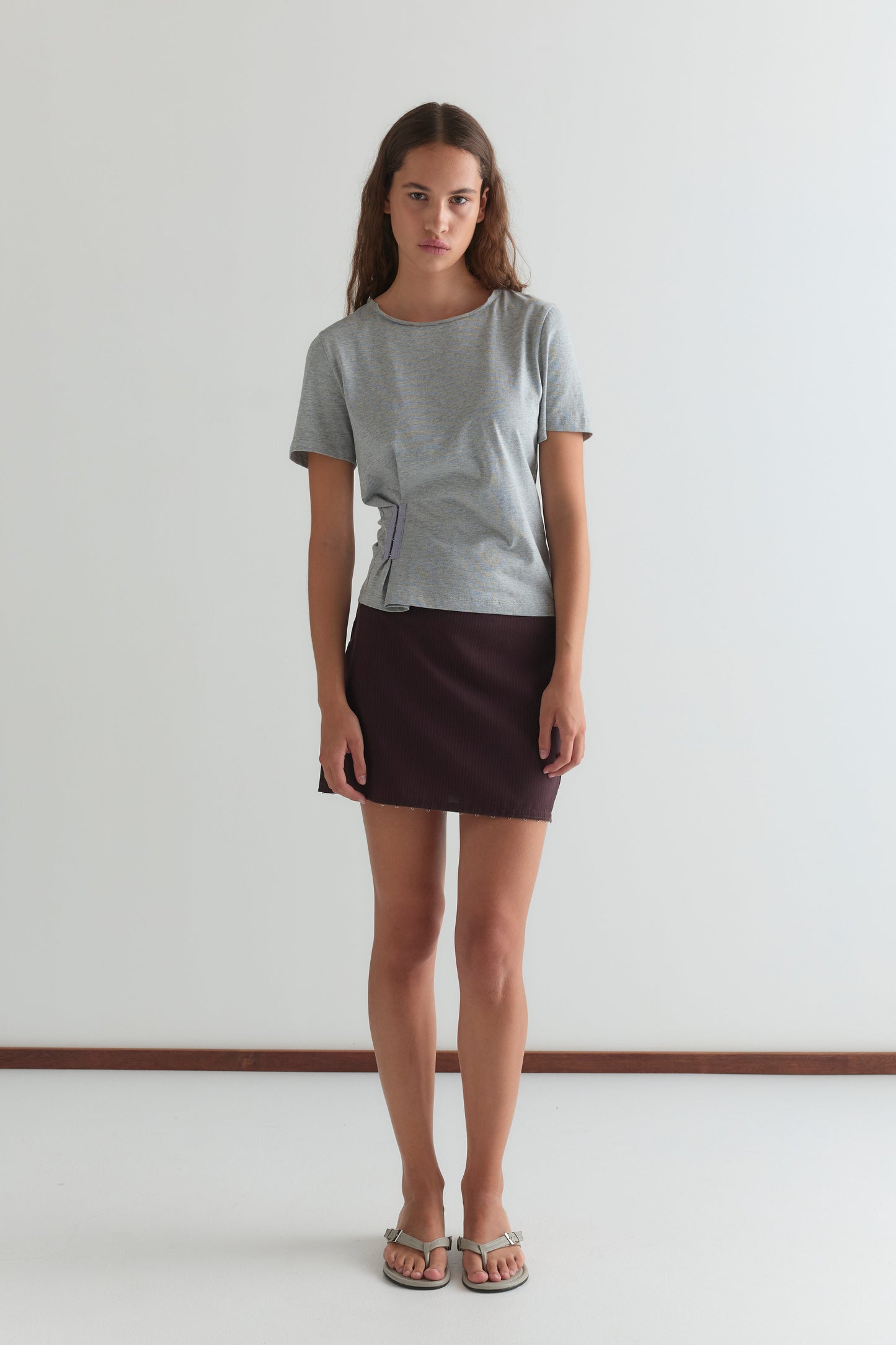 A young woman with long, wavy brown hair and a neutral expression stands indoors against a white wall, wearing a gray short-sleeve top, sandals, and the Eye Skirt - Burgundy Brown Stripe from Deiji Studios.