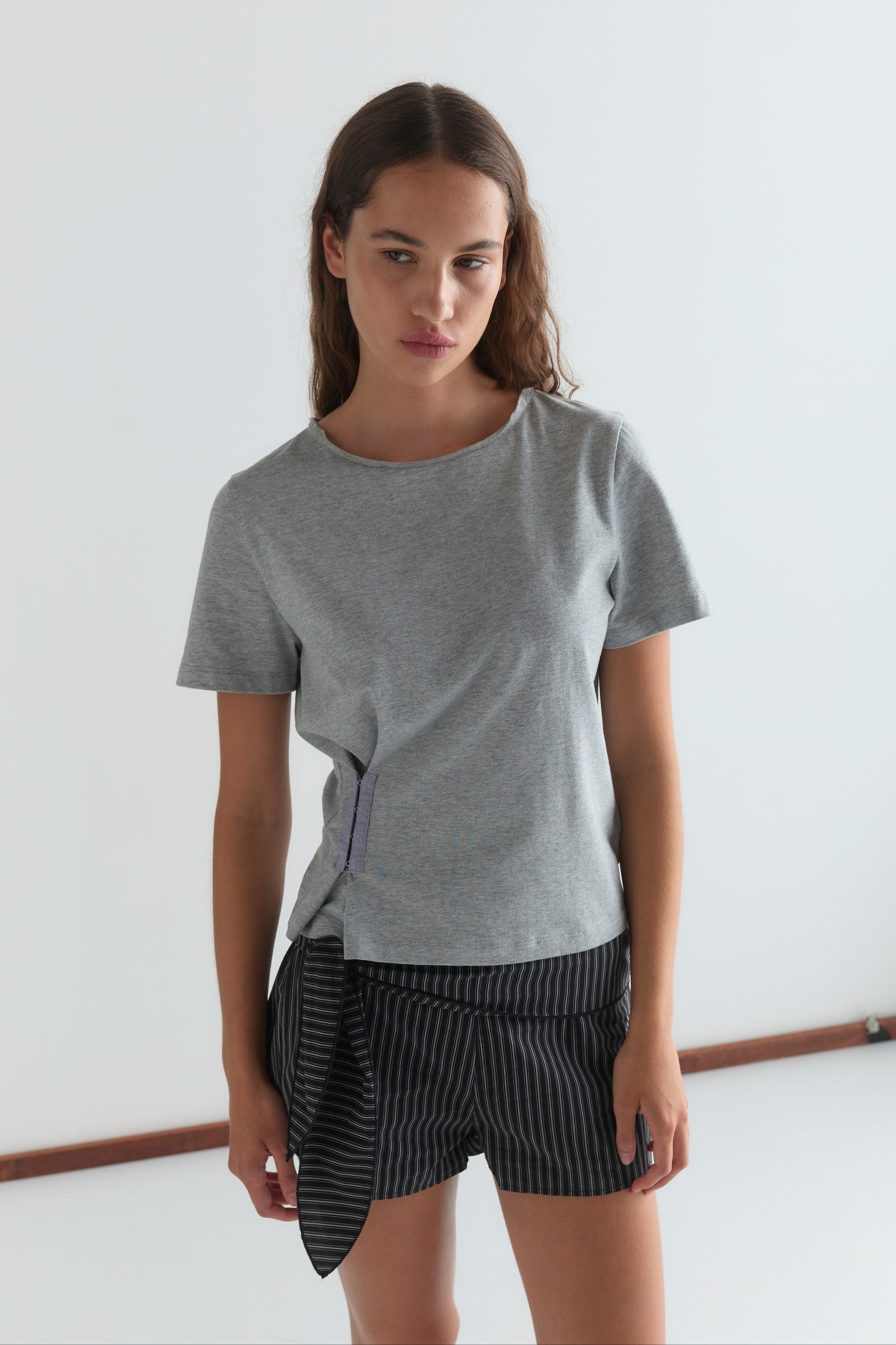 A woman with long brown hair stands against a white background, wearing a gray tee and the Belted Short - Mono Stripe by Deiji Studios, featuring black pinstripes and an asymmetric tie detail. She faces forward with a neutral expression.