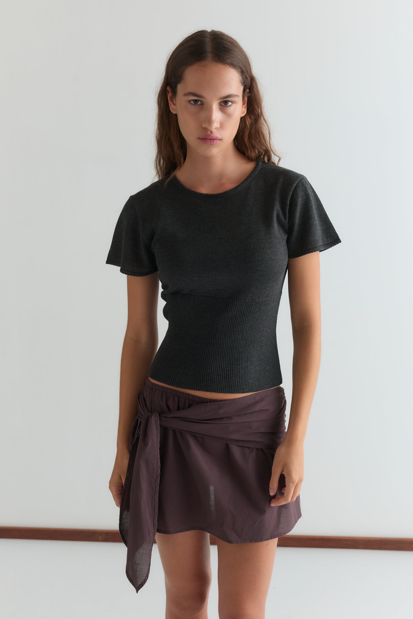 A woman with long brown hair wears a fitted black short-sleeve top and a wine-colored belted mini skirt (Belted Skirt - Wine), standing against a plain light background and looking directly at the camera with a neutral expression.