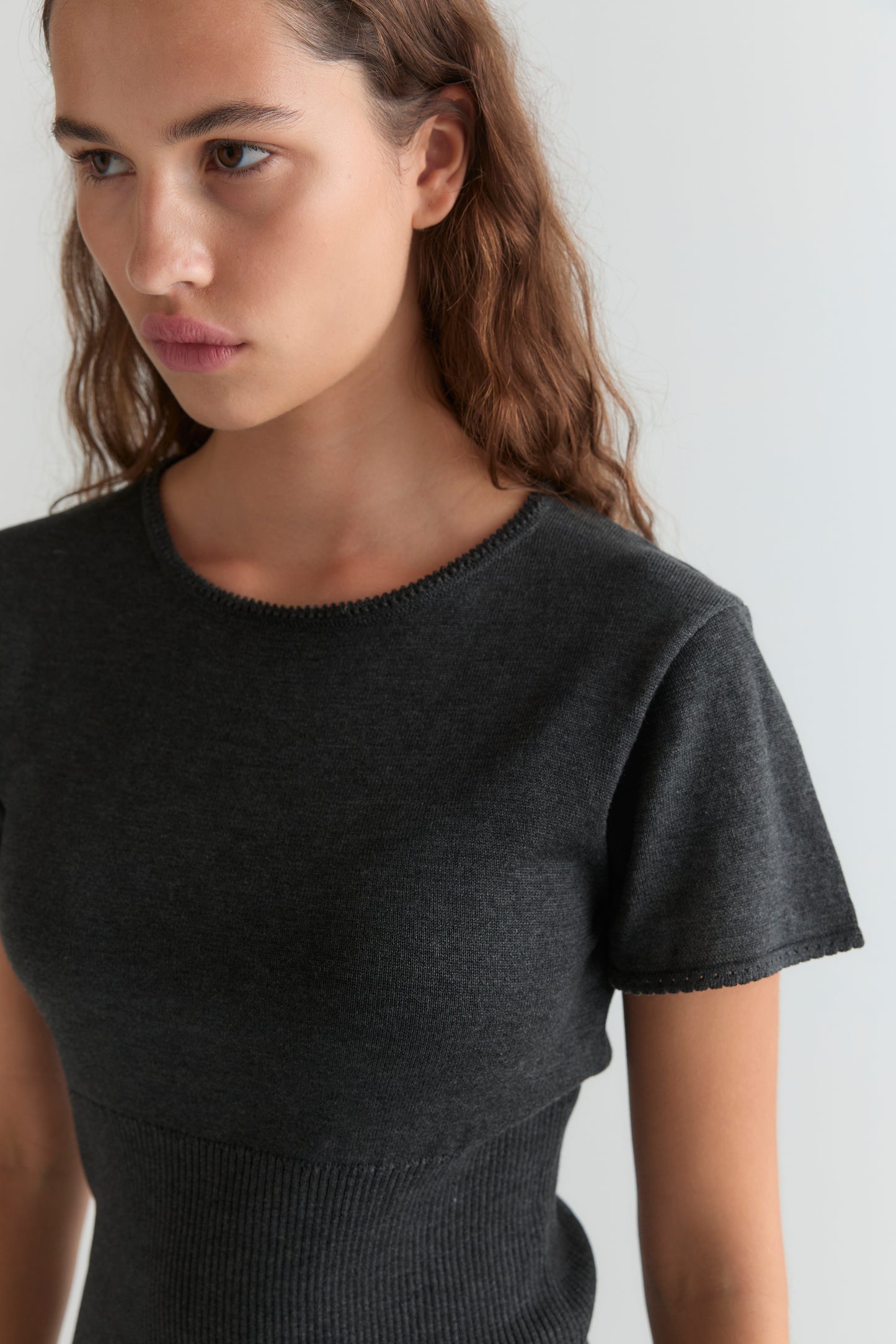 A young woman with wavy brown hair wears the Trim Knit Tee - Dark Grey, standing against a plain light background and looking slightly to the side with a neutral expression.