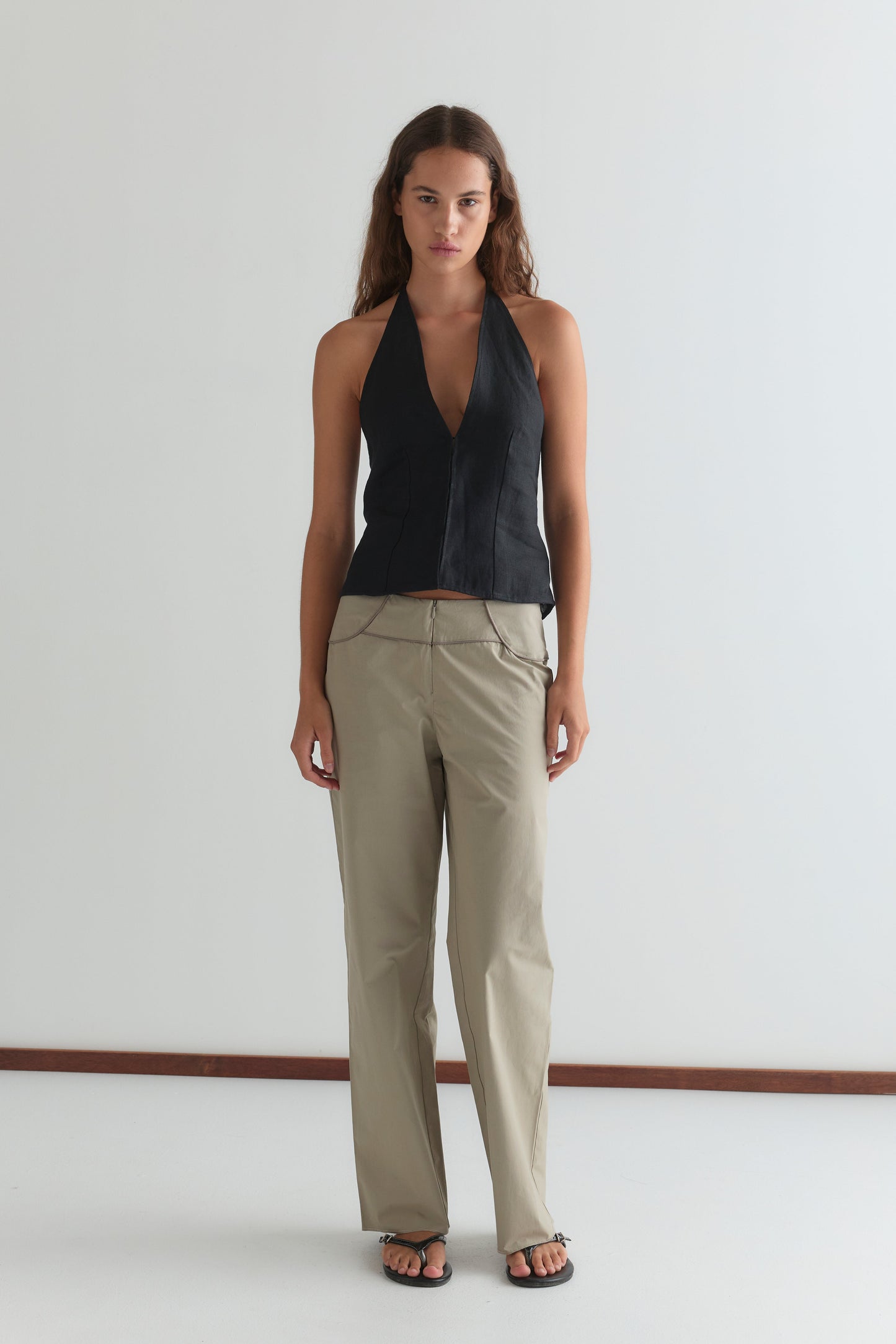 A woman with long brown hair and a neutral expression stands against a white wall, wearing the Close Halter Top - Black from Deiji Studios, beige zip-detail trousers, and black sandals.