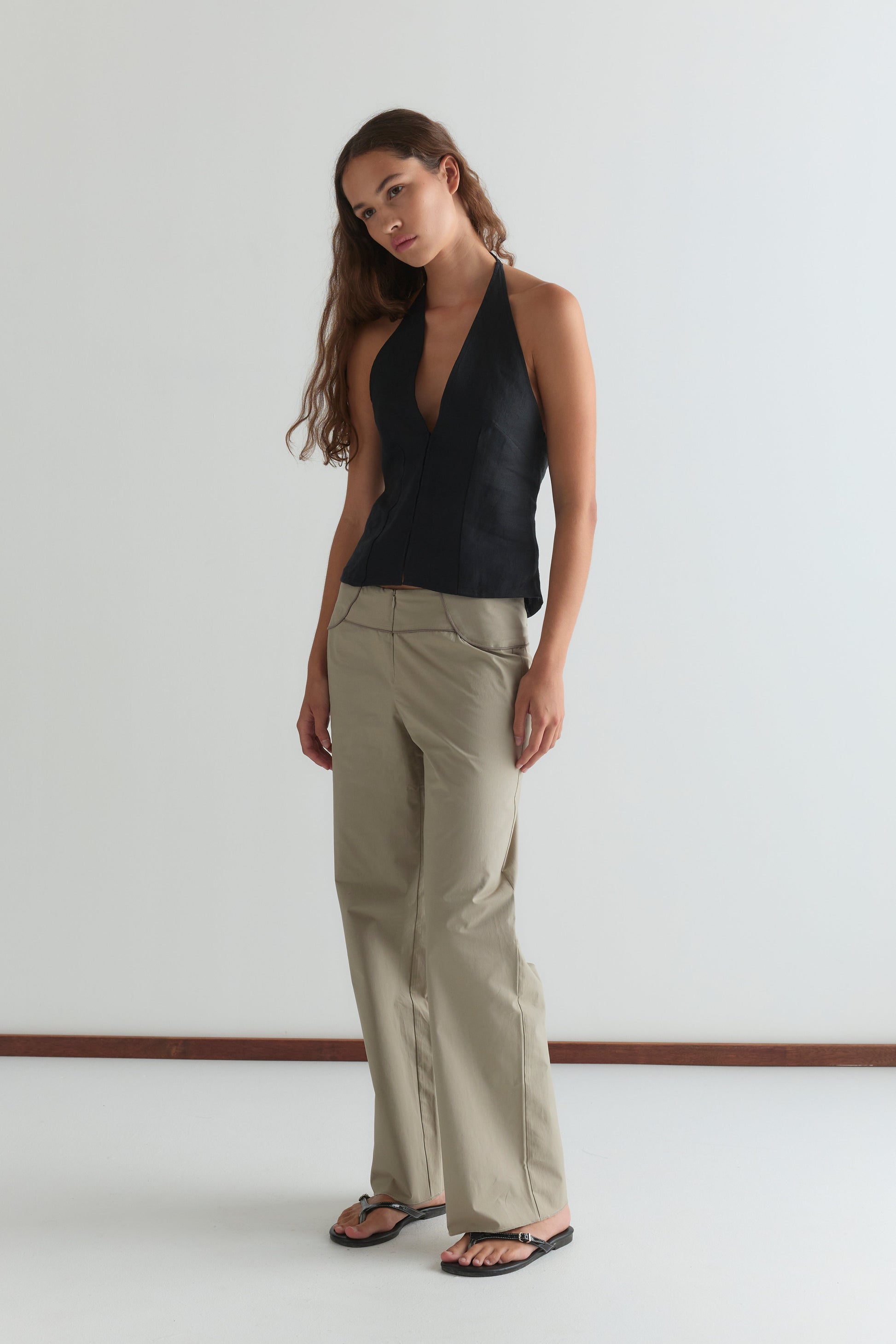 A woman with long brown hair wears the Close Halter Top - Black by Deiji Studios, paired with beige wide-leg pants and black sandals, standing against a plain white background.