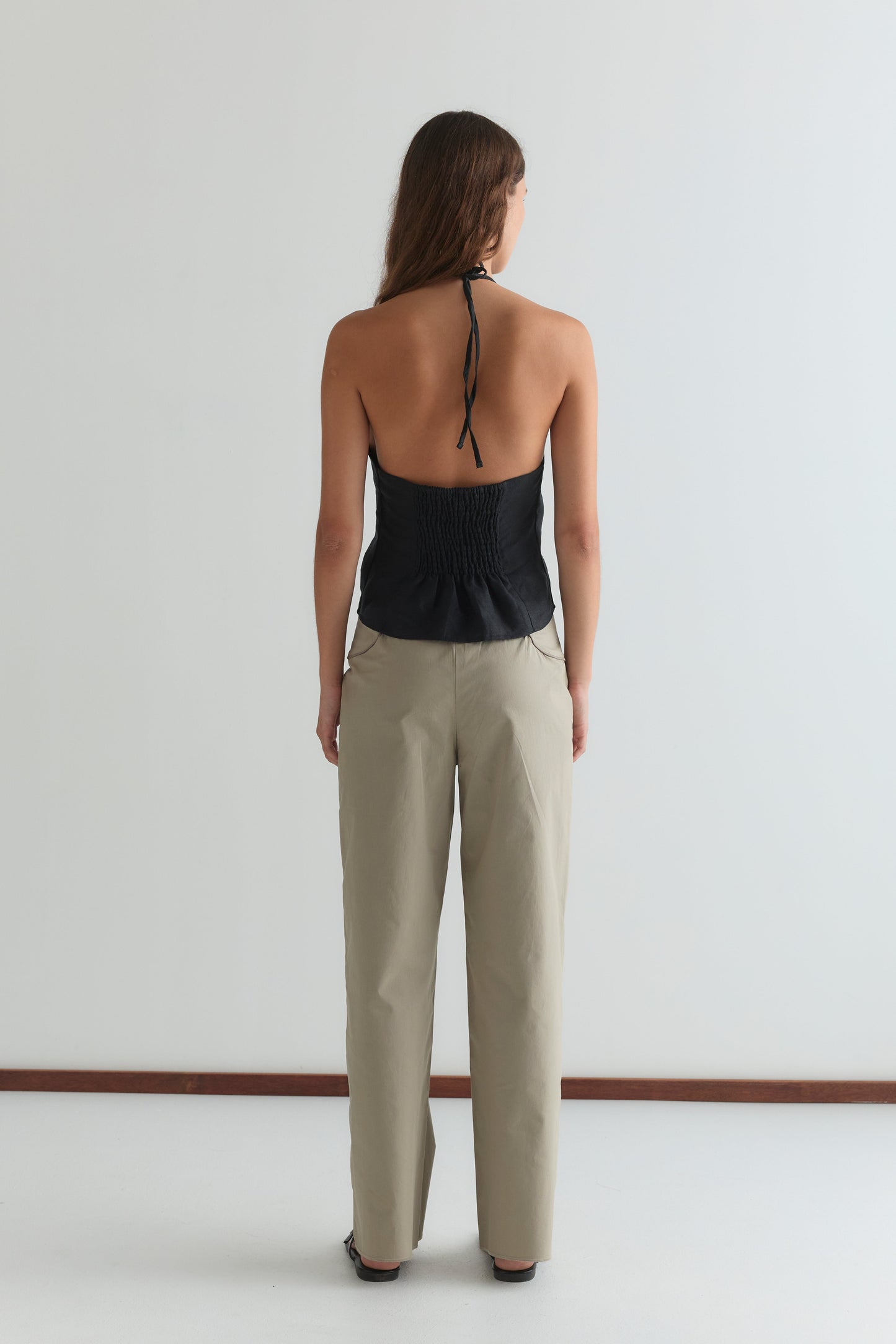 A woman faces away from the camera, wearing the Close Halter Top - Black by Deiji Studios with an open back and beige pants against a plain white background.