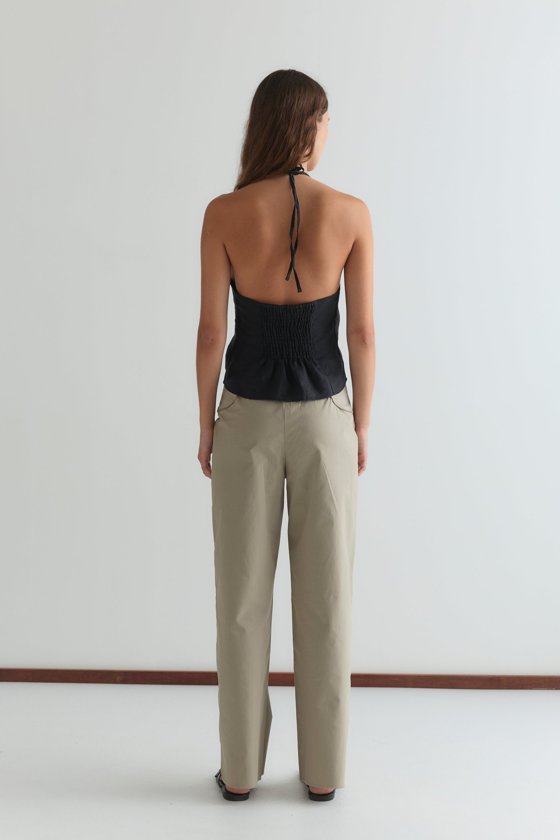 A woman faces away from the camera, wearing the Close Halter Top - Black by Deiji Studios with an open back and beige pants against a plain white background.