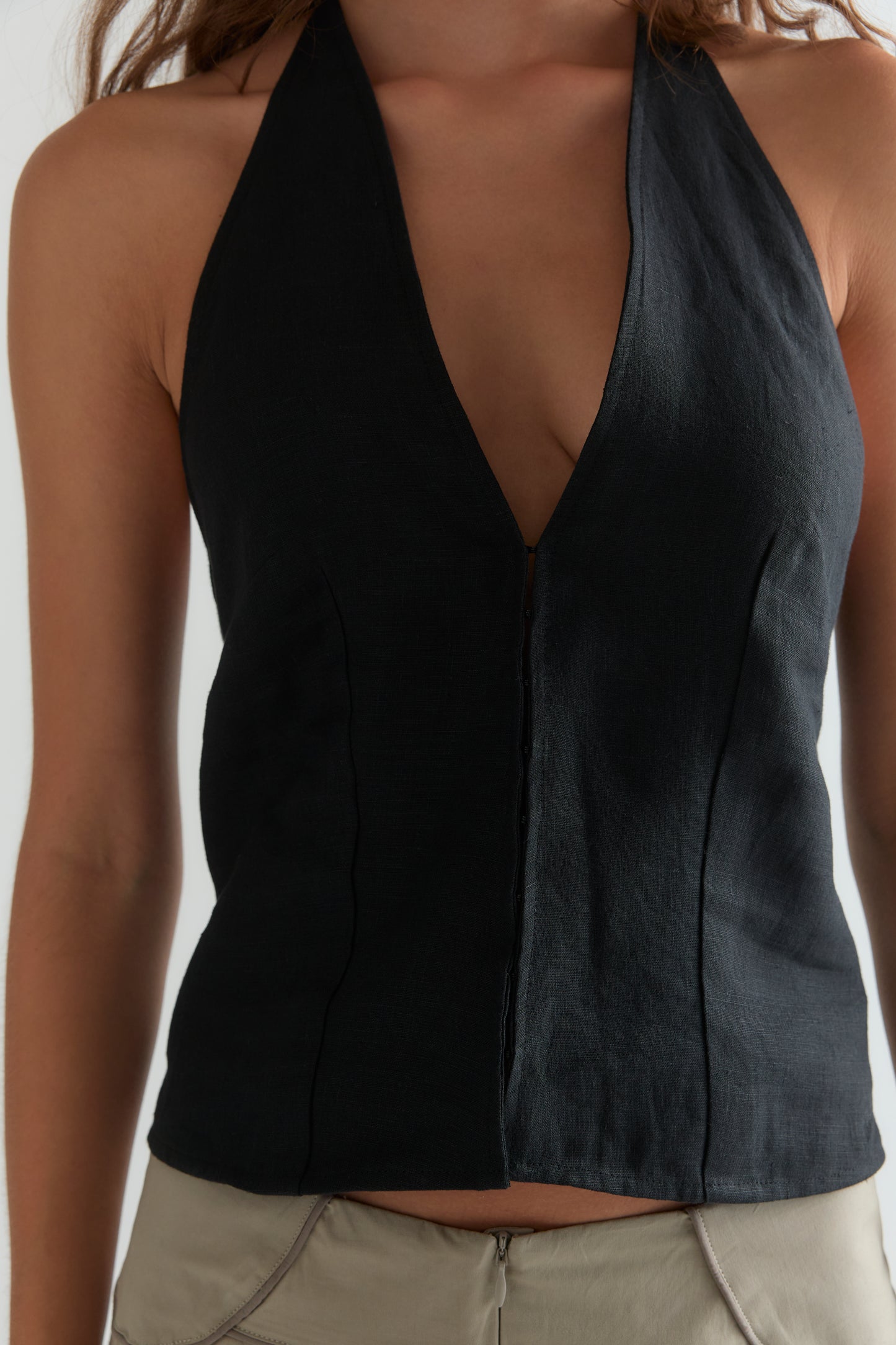 A person wearing the Close Halter Top - Black by Deiji Studios, a sleeveless halter style with a deep V-neckline, paired with beige pants, shown from shoulders to waist.