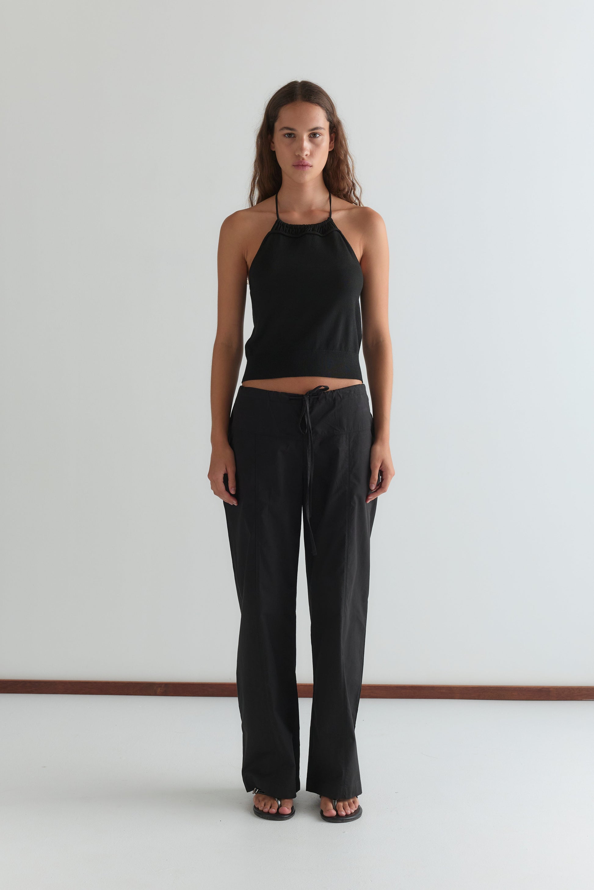A woman with long wavy hair and a neutral expression stands against a white wall, wearing black Seam Pants, a black halter-neck top, and black sandals, with her arms relaxed at her sides.