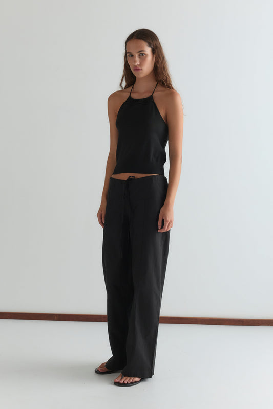A woman with long, wavy brown hair and a neutral expression stands indoors against a plain white wall, wearing Deiji Studios' Drawcord Knit Top - Black, loose black pants, and black sandals.
