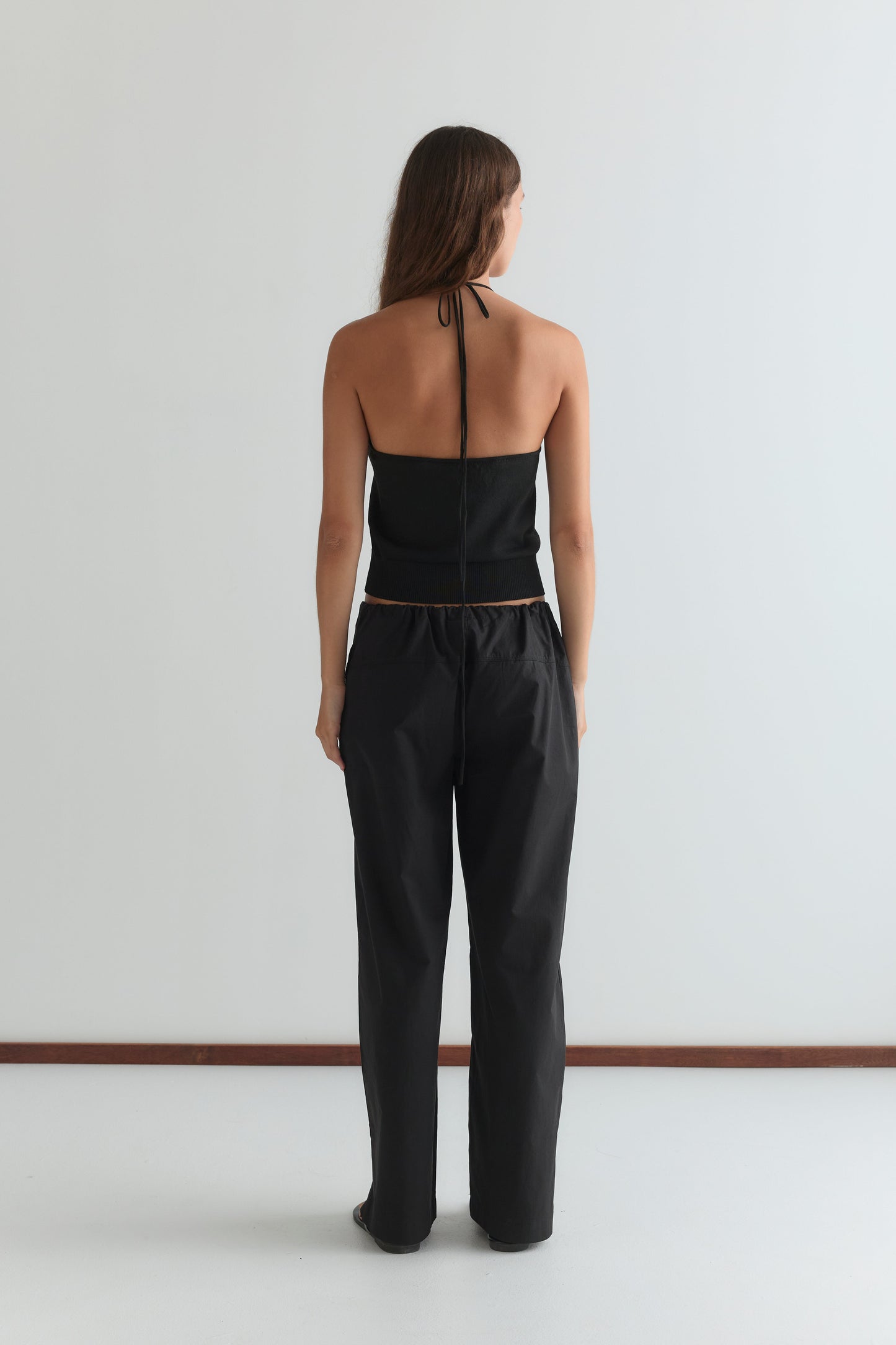 A person with long hair, facing away, wears the Seam Pant - Black and a black halter-top, standing against a plain white wall and floor.