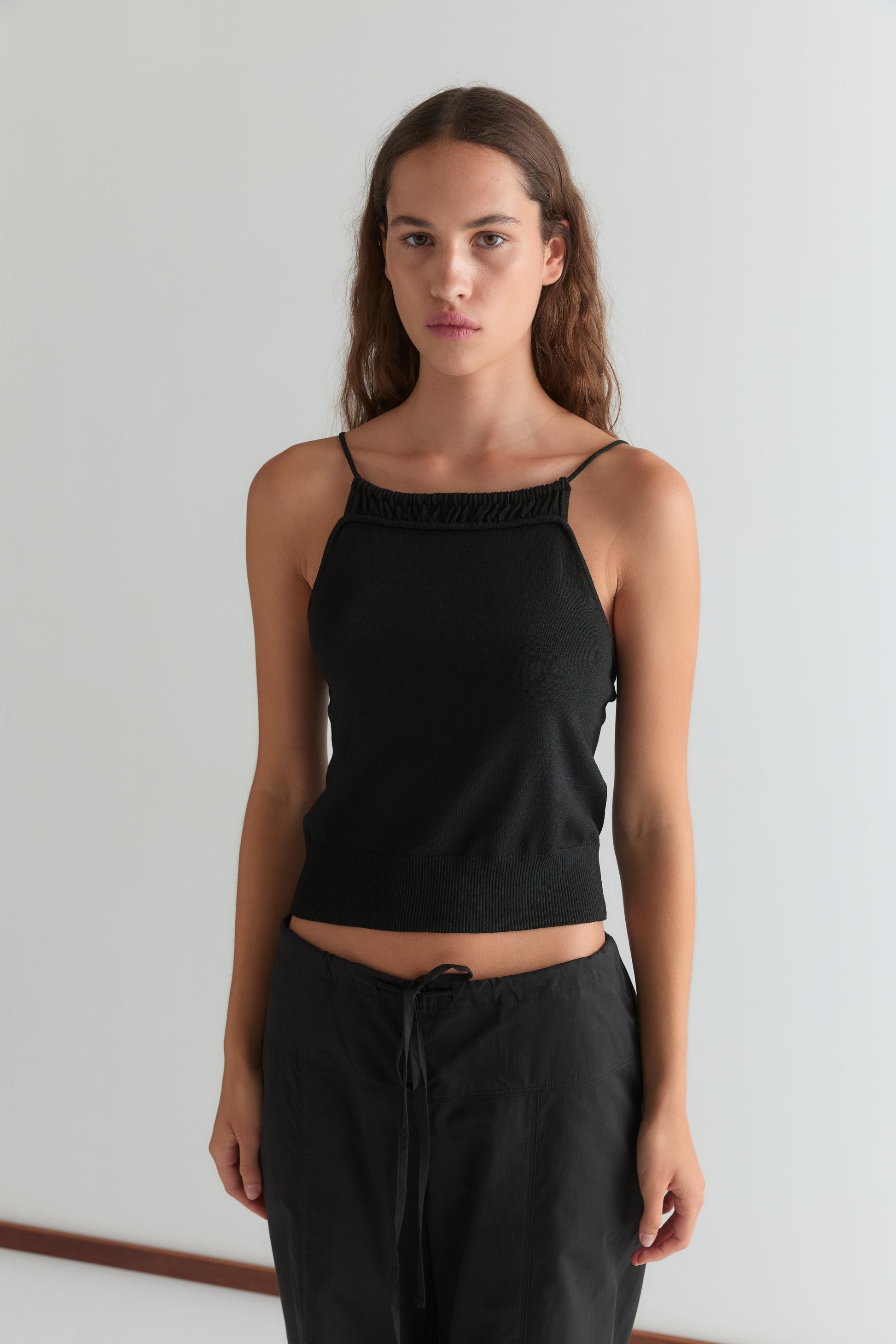 A person with long brown hair stands against a white wall, wearing the black Seam Pant for a comfortable fit, paired with a black sleeveless top, looking directly at the camera with a neutral expression.
