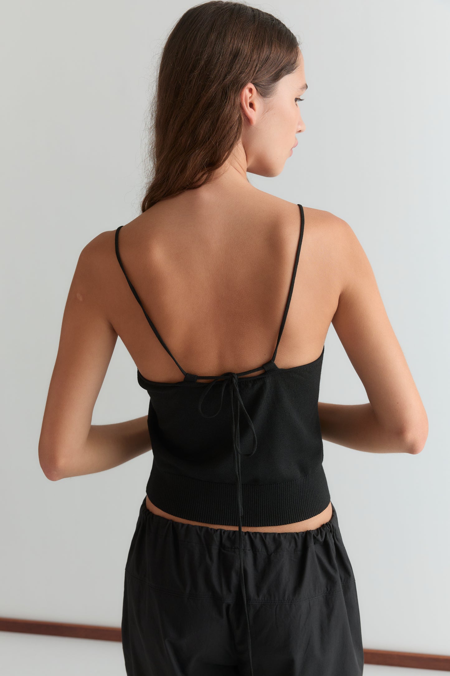 A woman with long brown hair faces away, wearing the Deiji Studios Drawcord Knit Top - Black with an adjustable ruched neckline and open back, paired with black pants against a plain light background.