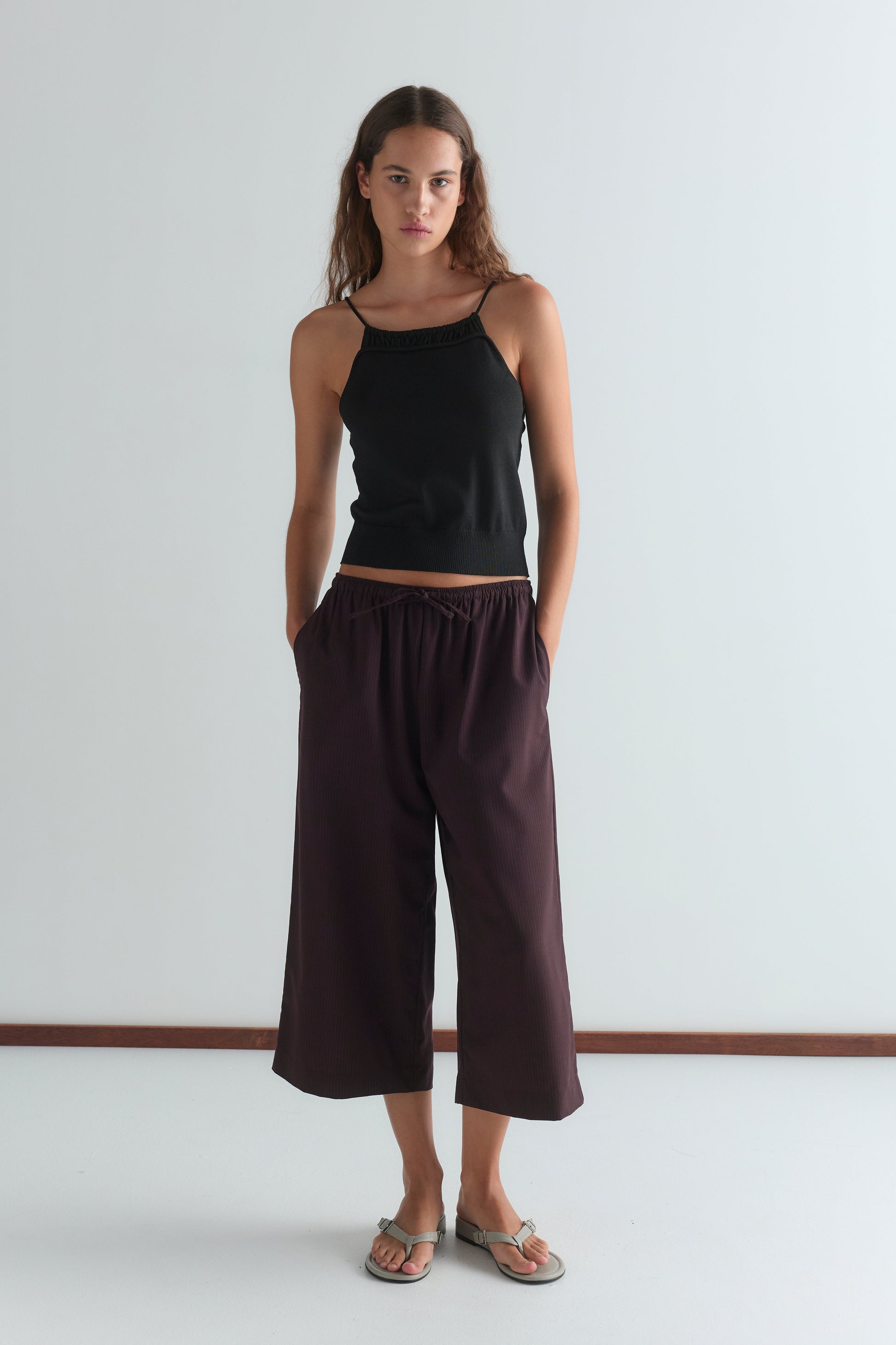 A woman stands against a plain white wall wearing a black camisole, Drawcord Culotte - Burgundy Brown Stripe by Deiji Studios, and sandals. Her long hair is down and her hands rest in her pockets.