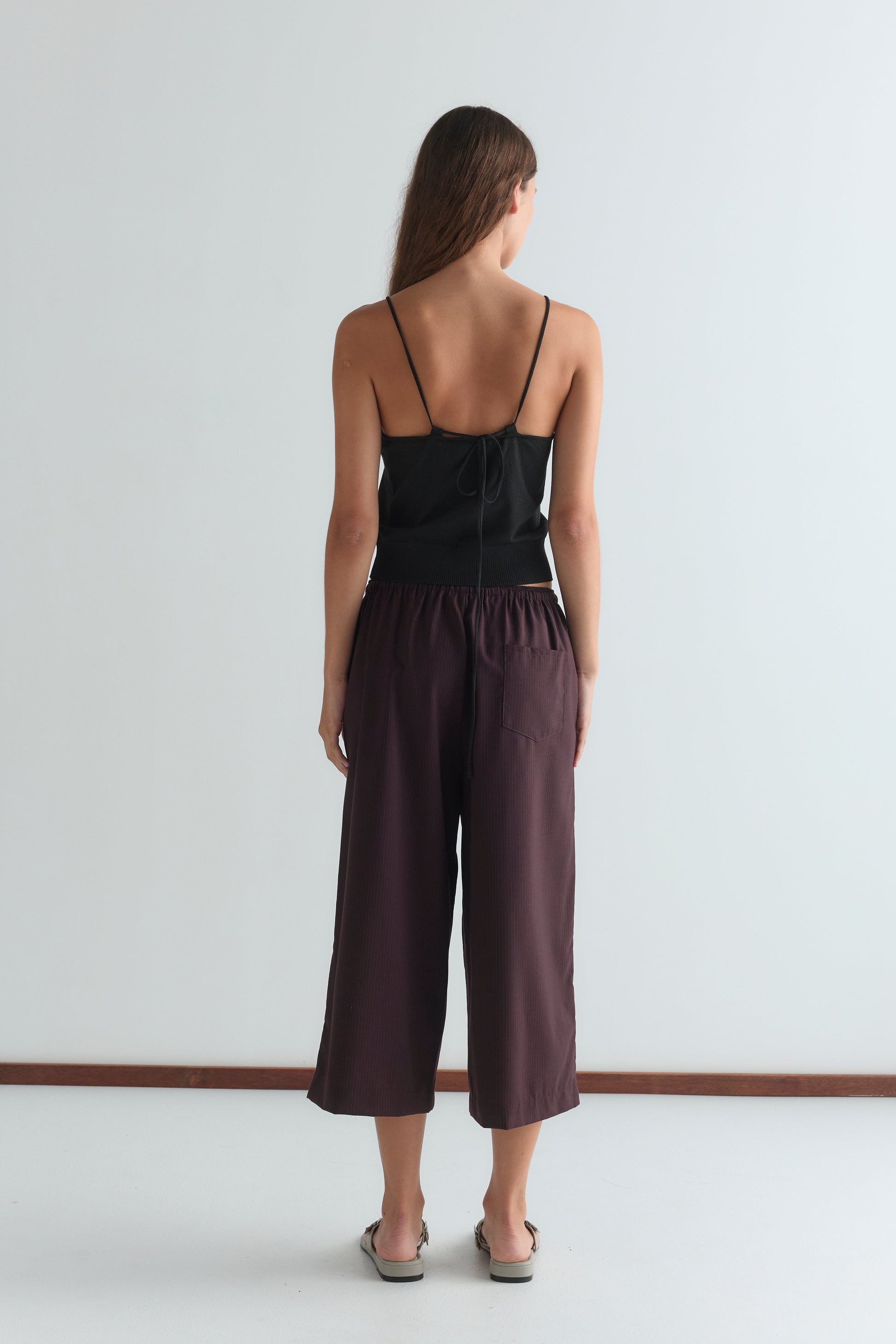 A woman with long brown hair faces away from the camera, wearing a relaxed black spaghetti strap top and Drawcord Culotte - Burgundy Brown Stripe pants in a minimalist room with a white wall and brown floor trim.