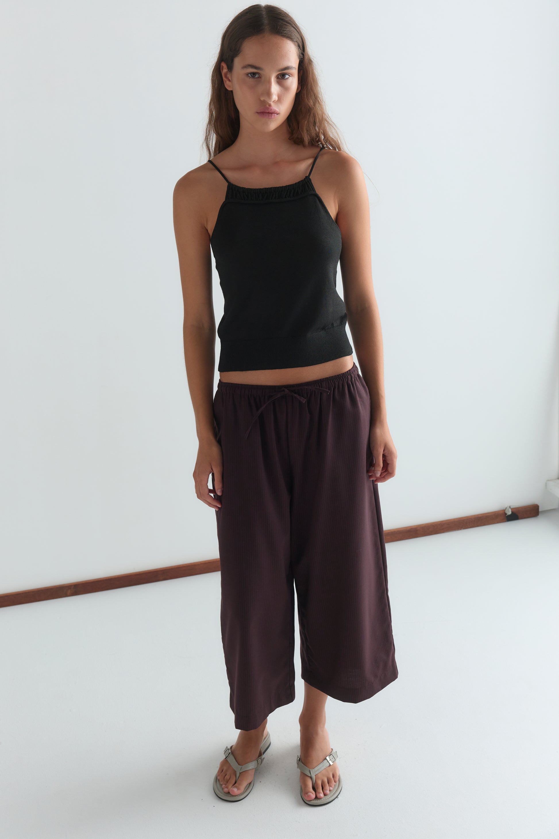A woman with long brown hair wears a black sleeveless top, Drawcord Culotte - Burgundy Brown Stripe by Deiji Studios, and sandals, standing against a plain white background.