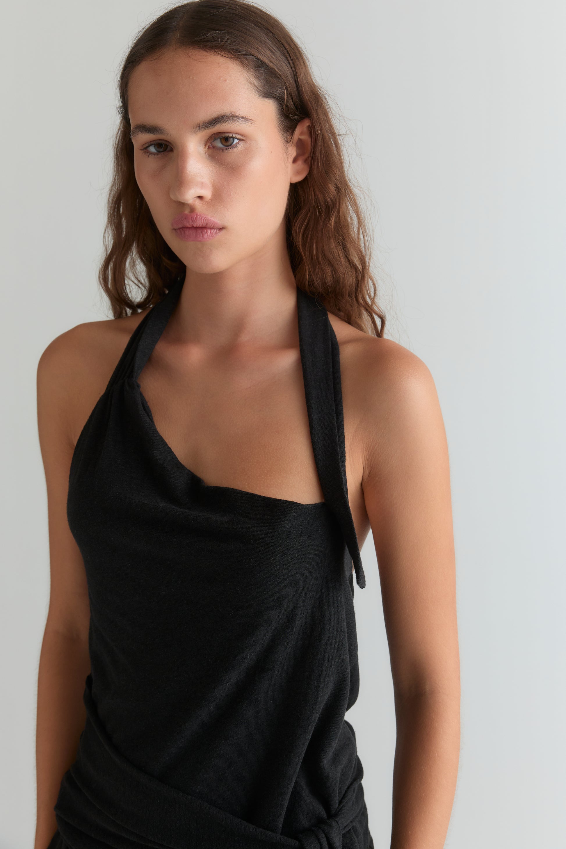 A woman with long brown hair wears the Drape Halter - Black and stands against a plain light background, looking at the camera with a neutral expression.