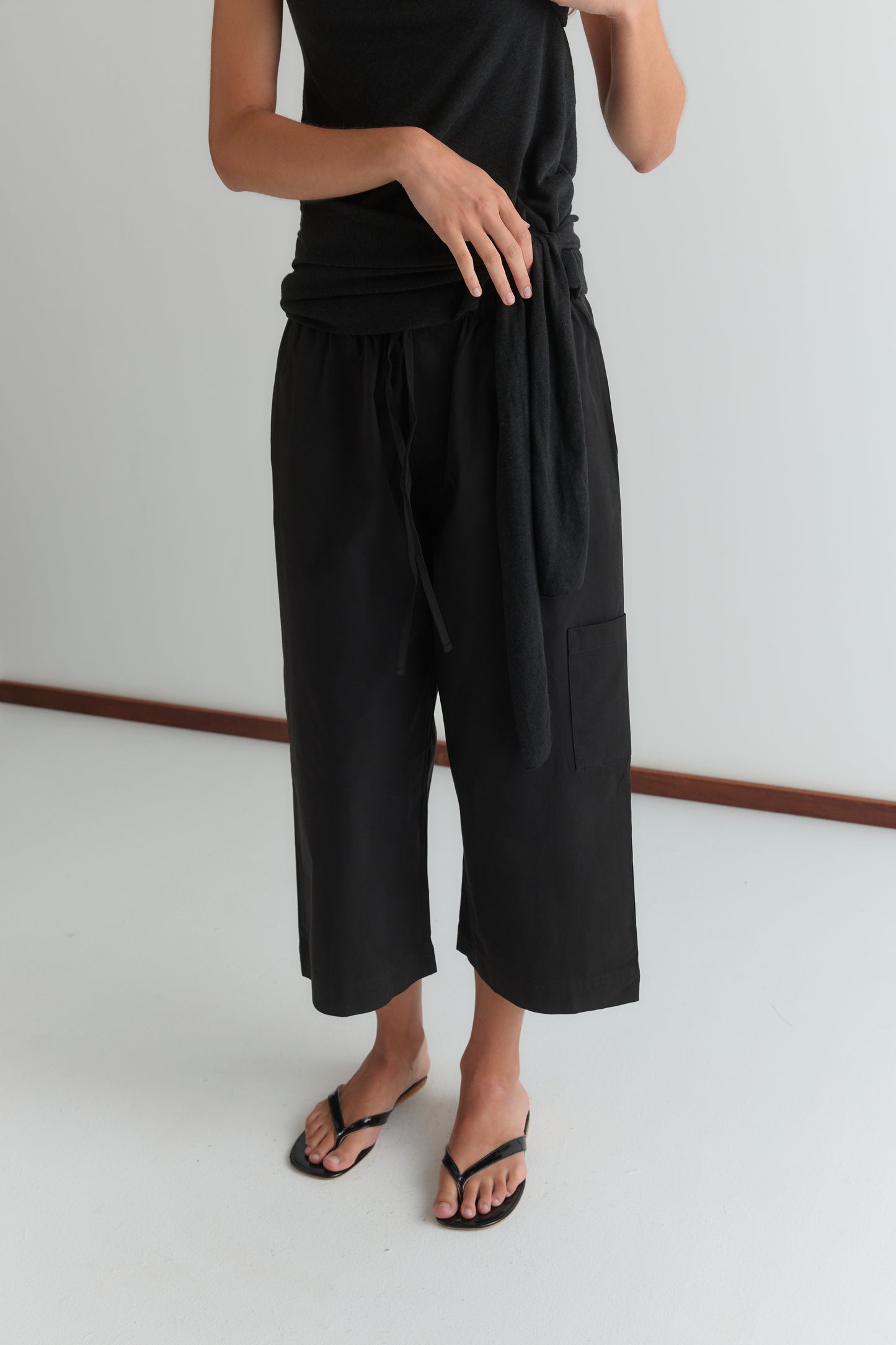A person wears a Deiji Studios black sleeveless top with the Mid Pocket Short - Black, styled as relaxed 3/4 wide-leg cropped pants, and black flip-flops, standing on a white floor against a plain light wall with their face not visible.