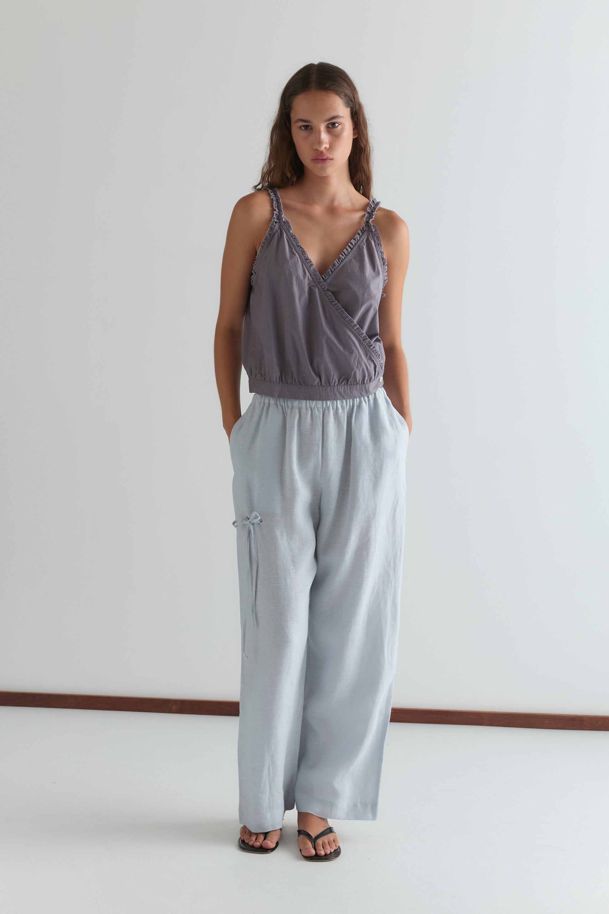 A woman stands against a plain white background wearing a loose, sleeveless gray wrap top and the Eyelet Pant - Soft Blue, featuring a relaxed ankle-length fit with tie details, hands in her pockets, and black sandals.
