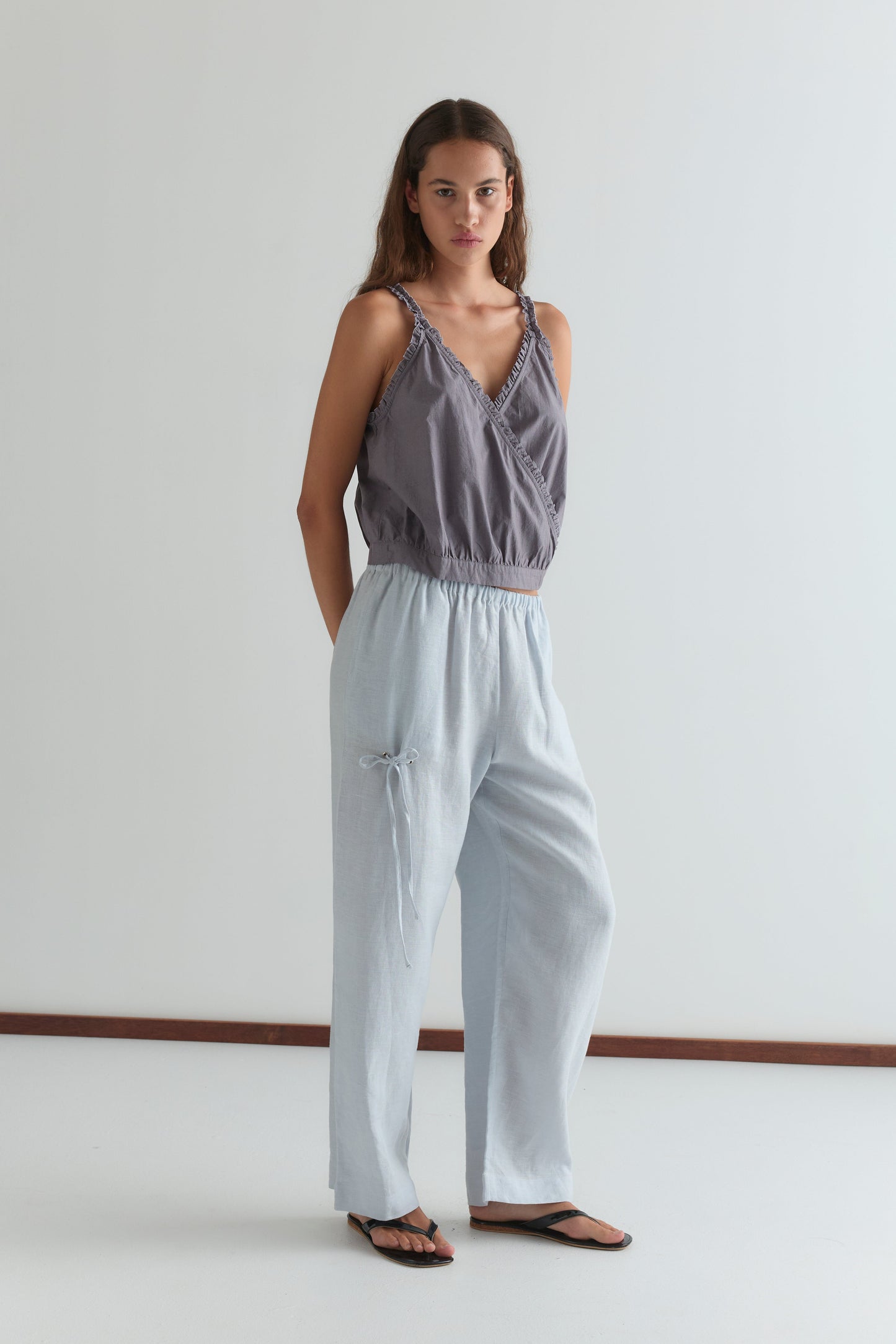A woman stands against a white wall, wearing Deiji Studios' Eyelet Pant in Soft Blue with a loose purple sleeveless top and black sandals. Her hands are in her pockets as she looks at the camera with a neutral expression.