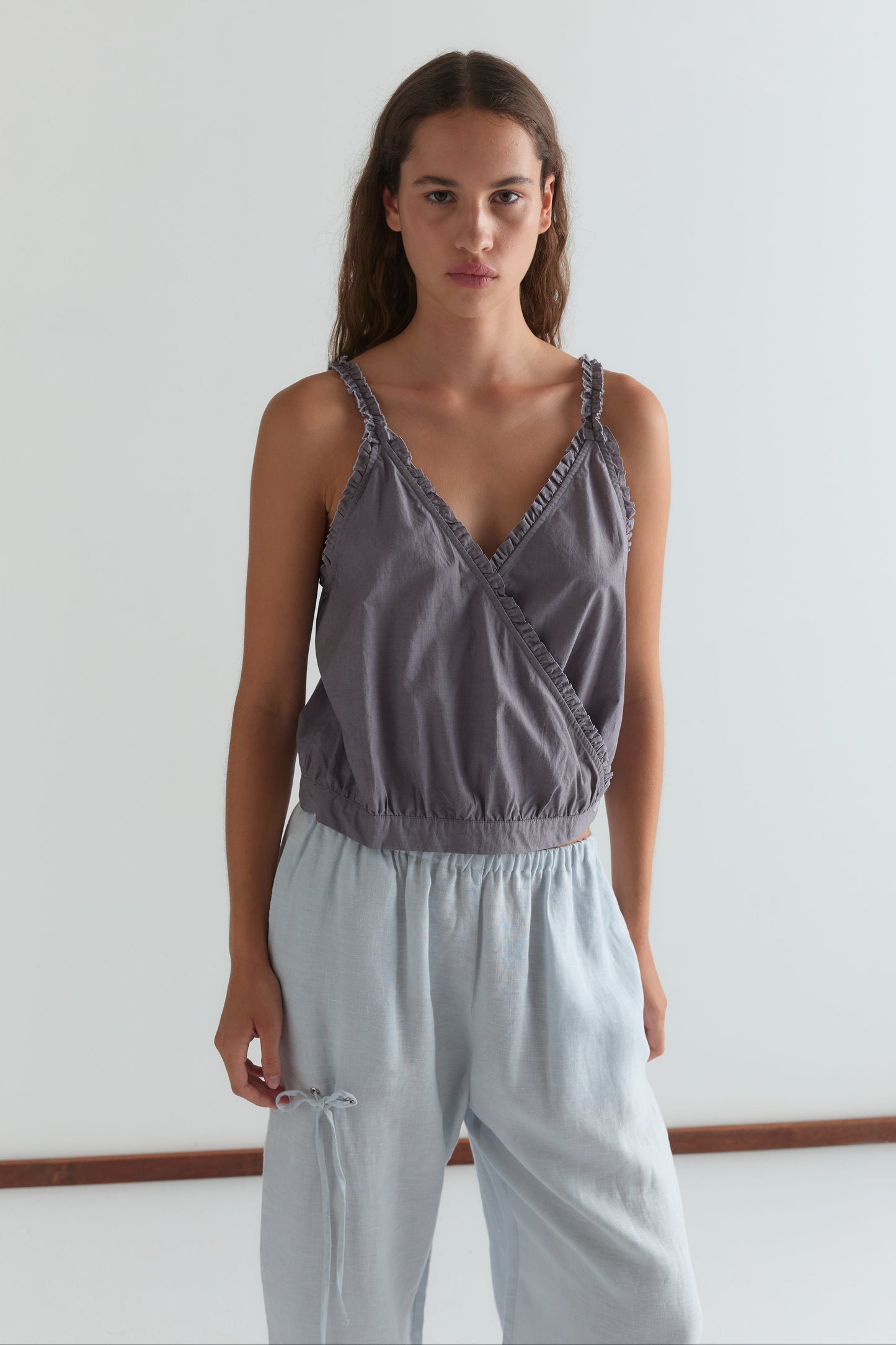 A woman with long brown hair stands against a white background, wearing a sleeveless ruffled gray top and Deiji Studios Eyelet Pant in Soft Blue. She looks directly at the camera with a neutral expression.