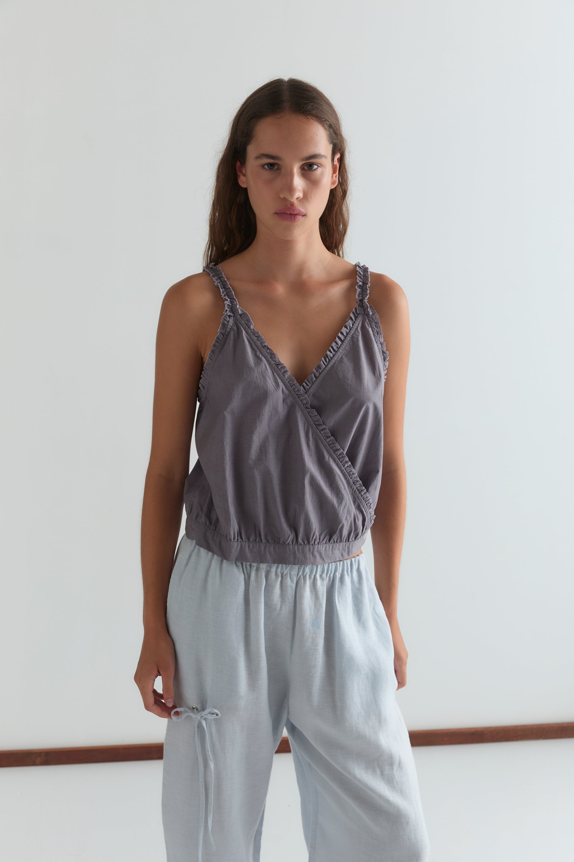 A woman with long brown hair stands against a plain white background, wearing the Overlie Top - Currant by Deiji Studios and loose light blue pants, looking directly at the camera with a neutral expression.