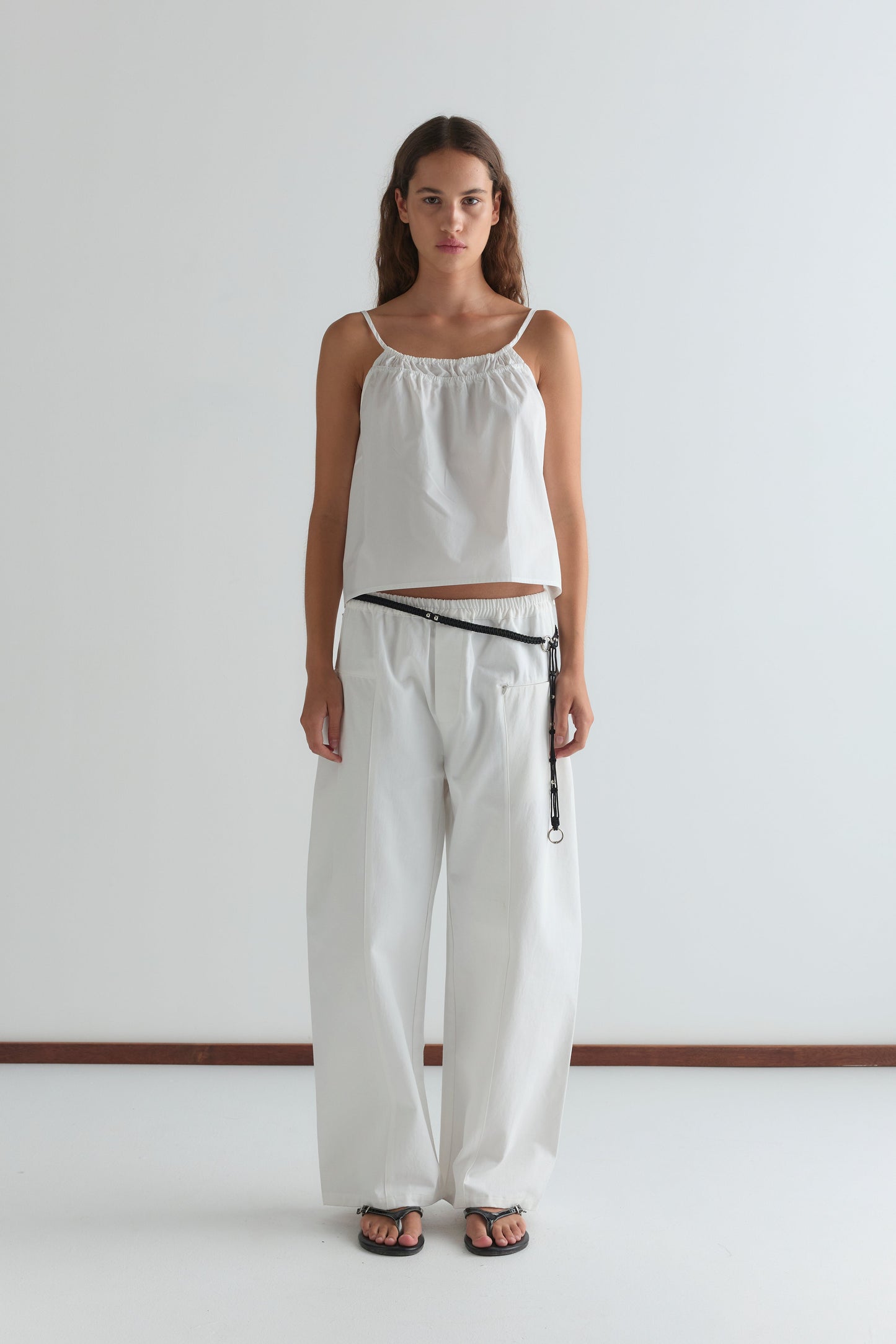 A woman stands against a white background wearing the Deiji Studios Double Layer Top - White with wide-legged white pants, a black belt featuring a hanging strap, and black sandals. She faces forward with a neutral expression.