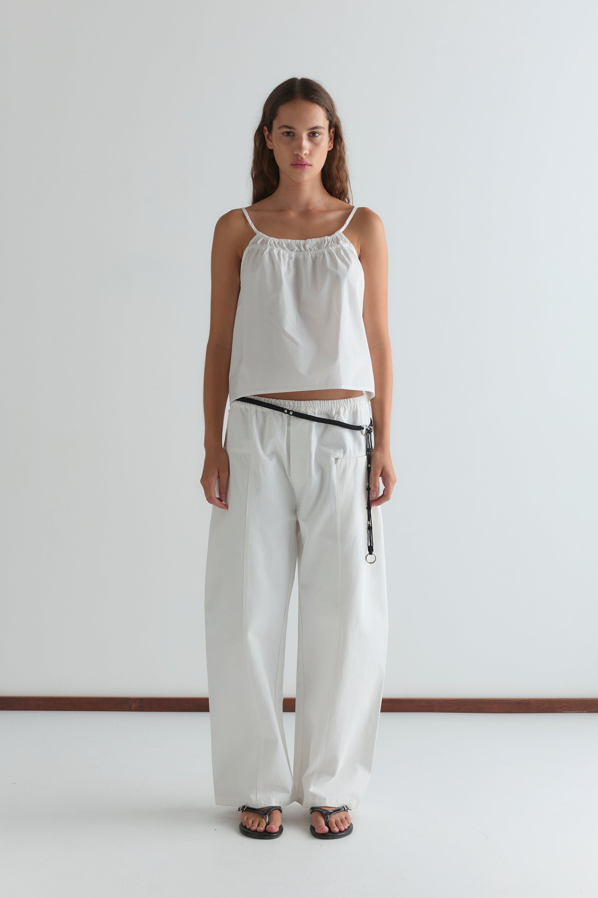 A woman stands against a white background wearing the Deiji Studios Double Layer Top - White with wide-legged white pants, a black belt featuring a hanging strap, and black sandals. She faces forward with a neutral expression.
