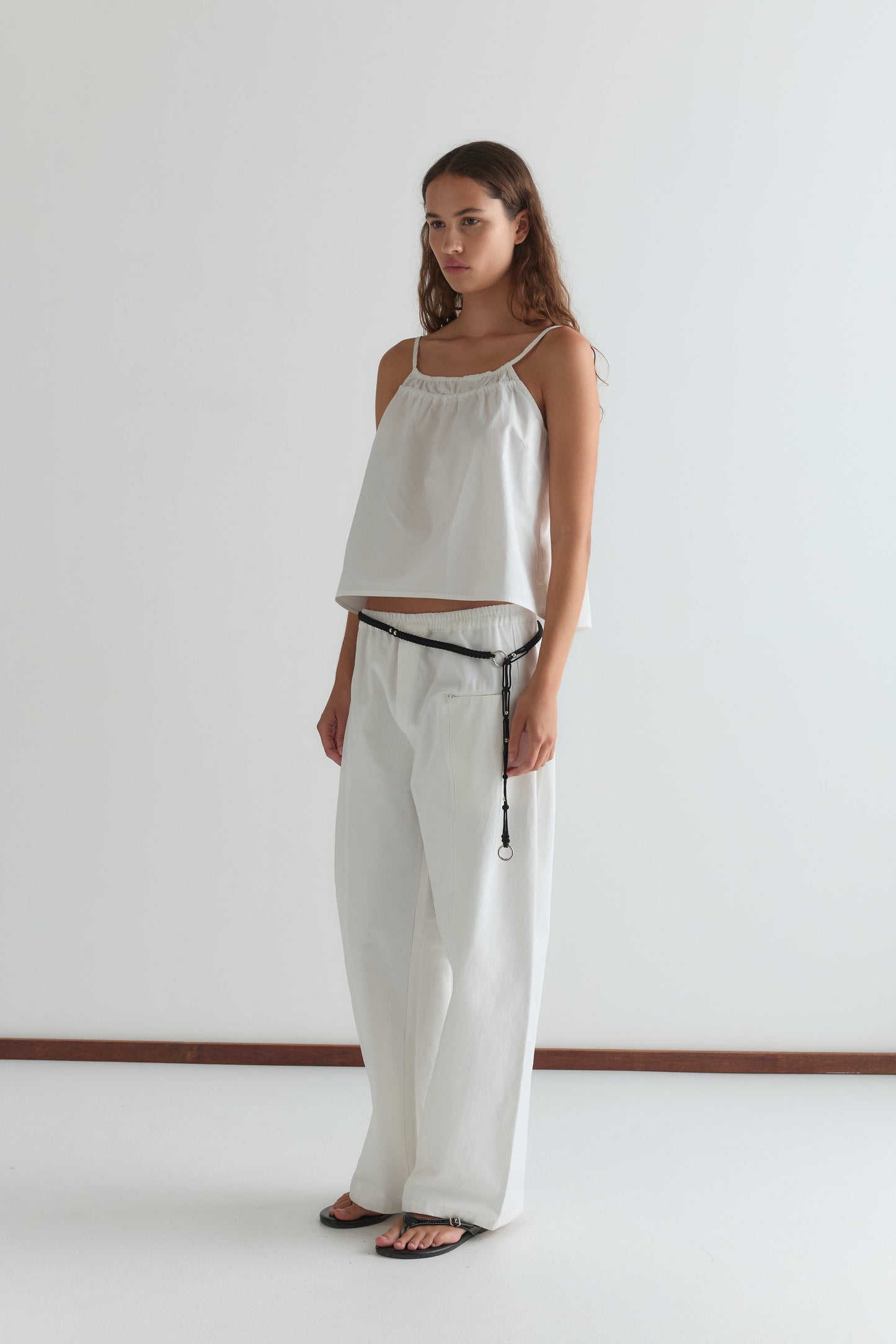 A woman stands in a minimal studio wearing Deiji Studios’ Double Layer Top - White, wide-leg white pants, a black cord belt, and black sandals. She has long brown hair and a neutral expression.