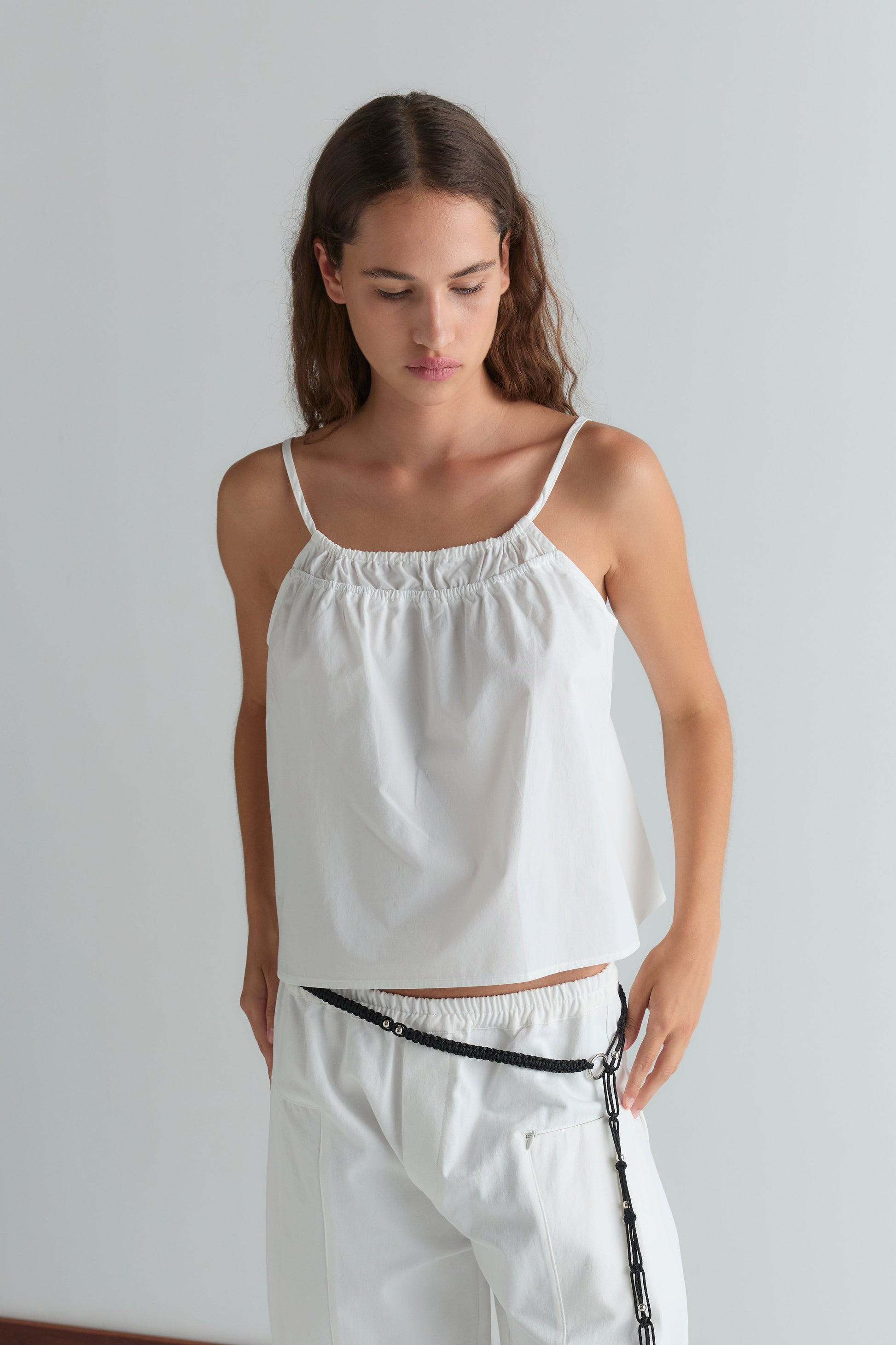 A woman wears the Deiji Studios Double Layer Top - White with a gathered neckline and layered detail, paired with white wide-leg trousers and a black chain belt, set against a light grey background.