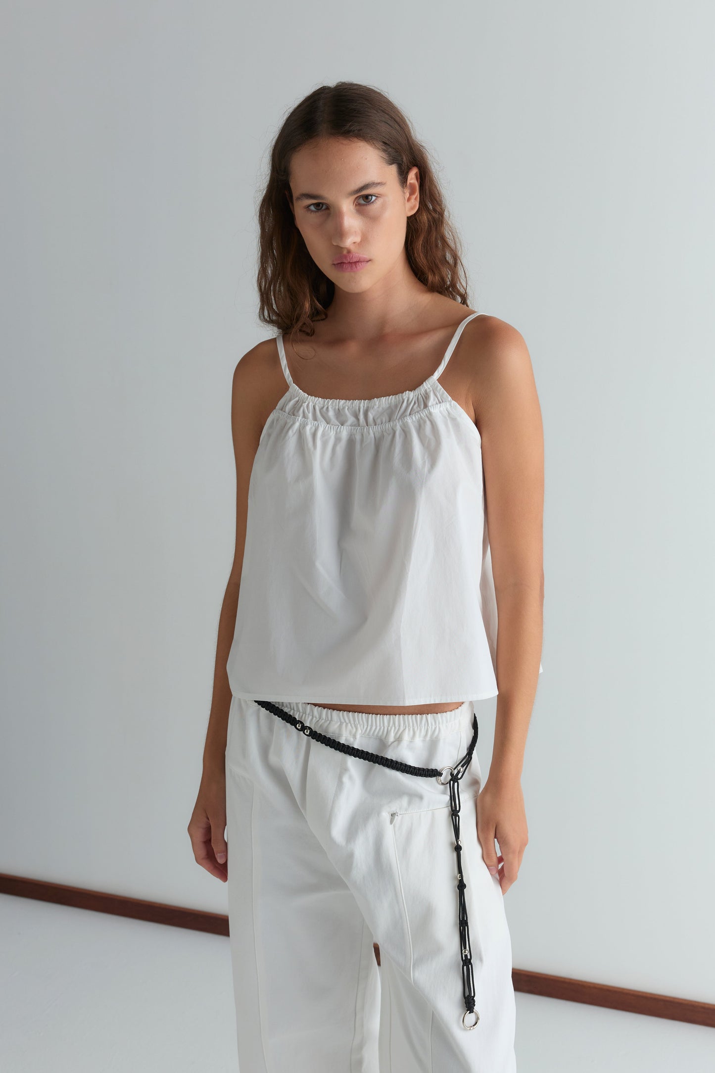 A woman stands against a plain white background, wearing the Double Layer Top - White and matching pants. She has a serious expression, with a black chain belt at her waist offering bold contrast.