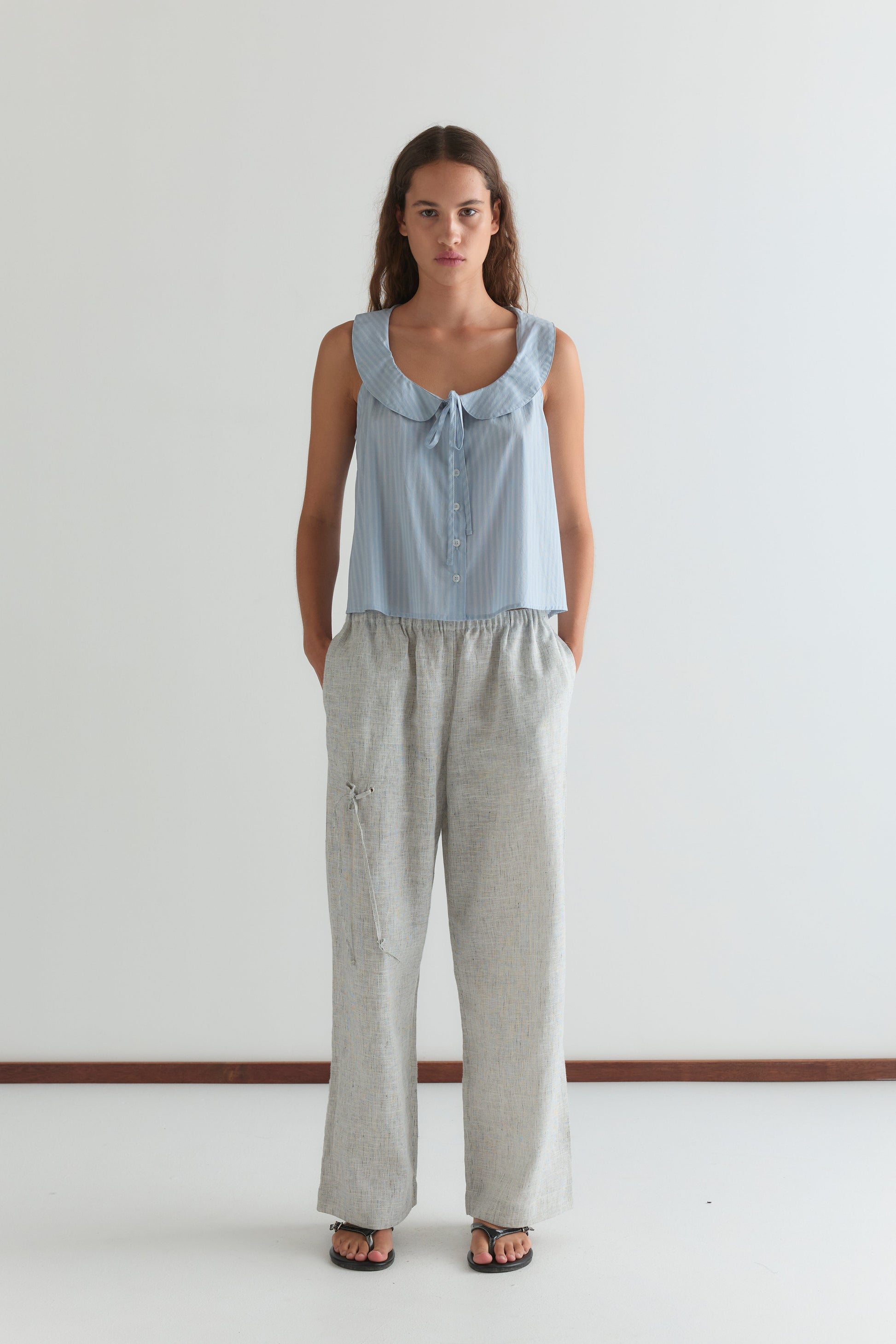 A woman stands against a plain white wall wearing a light blue sleeveless blouse, Eyelet Pant - Fine Stripe from Deiji Studios in gray, and black sandals. Her hands are in her pockets and she has a neutral expression.