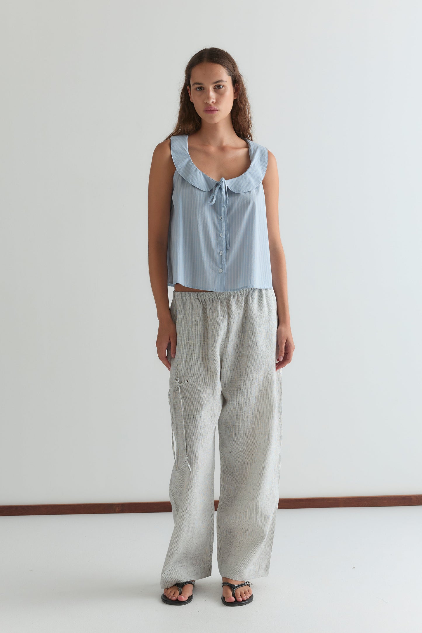 A woman stands indoors against a plain white wall, wearing a light blue sleeveless top with a ruffled neckline and front tie, Eyelet Pant - Fine Stripe by Deiji Studios in light gray, and black sandals.