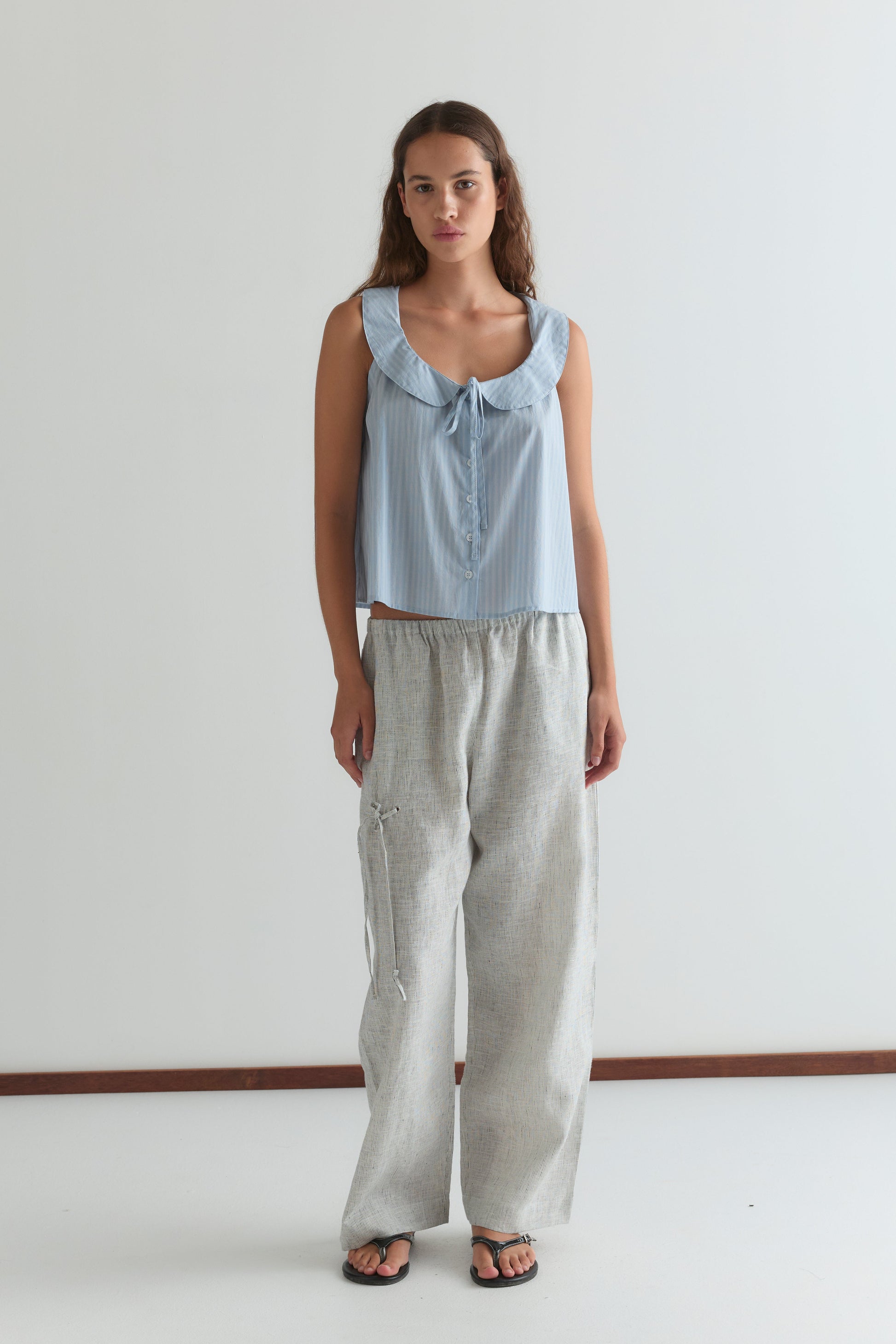 A woman stands indoors against a plain white wall, wearing a light blue sleeveless top with a ruffled neckline and front tie, Eyelet Pant - Fine Stripe by Deiji Studios in light gray, and black sandals.