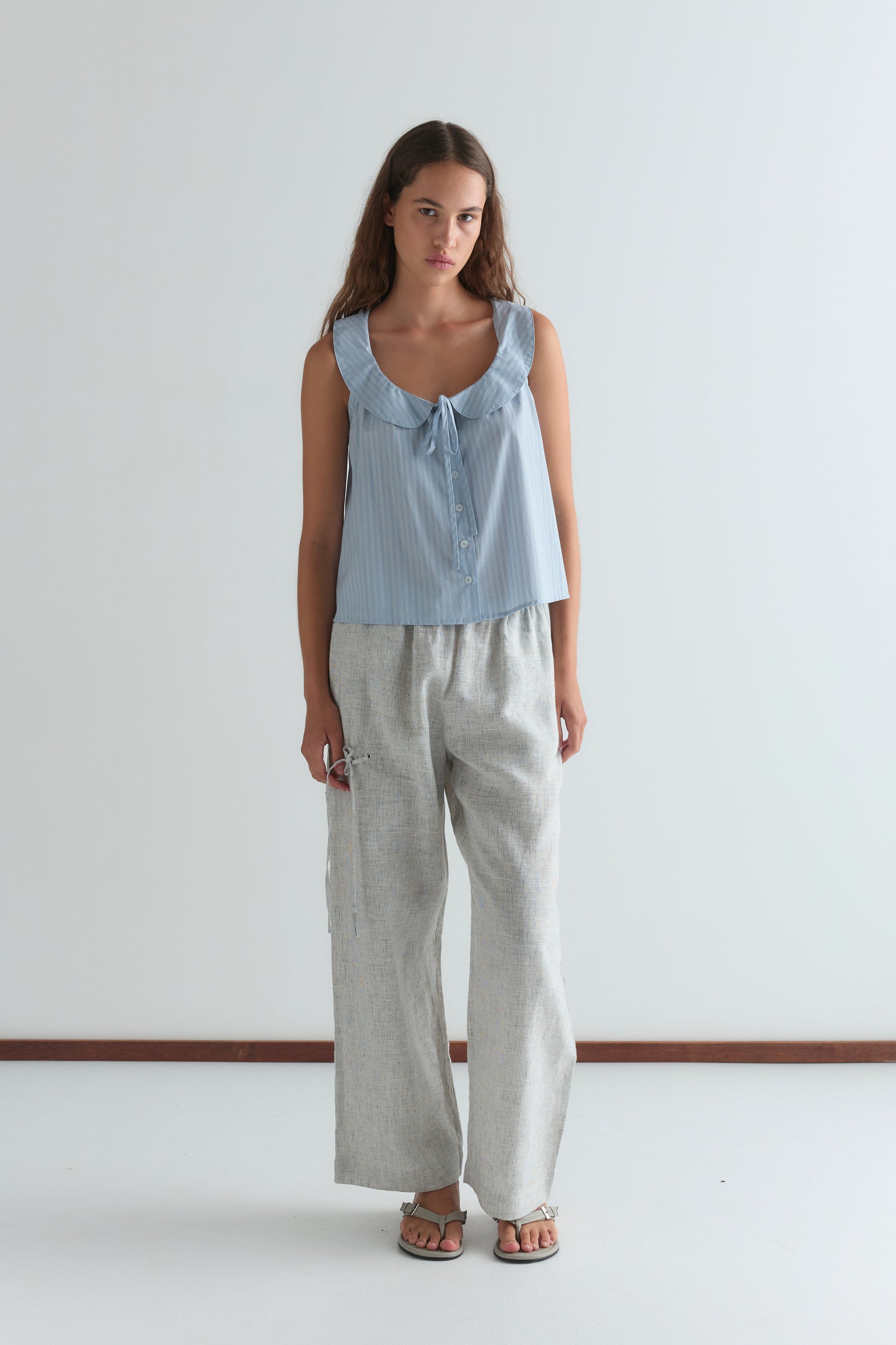 A woman with long brown hair stands indoors against a plain white wall, wearing the Round Collar Top - Lake Blue Stripe by Deiji Studios, featuring a ruffled neckline, paired with loose light gray patterned pants and sandals.