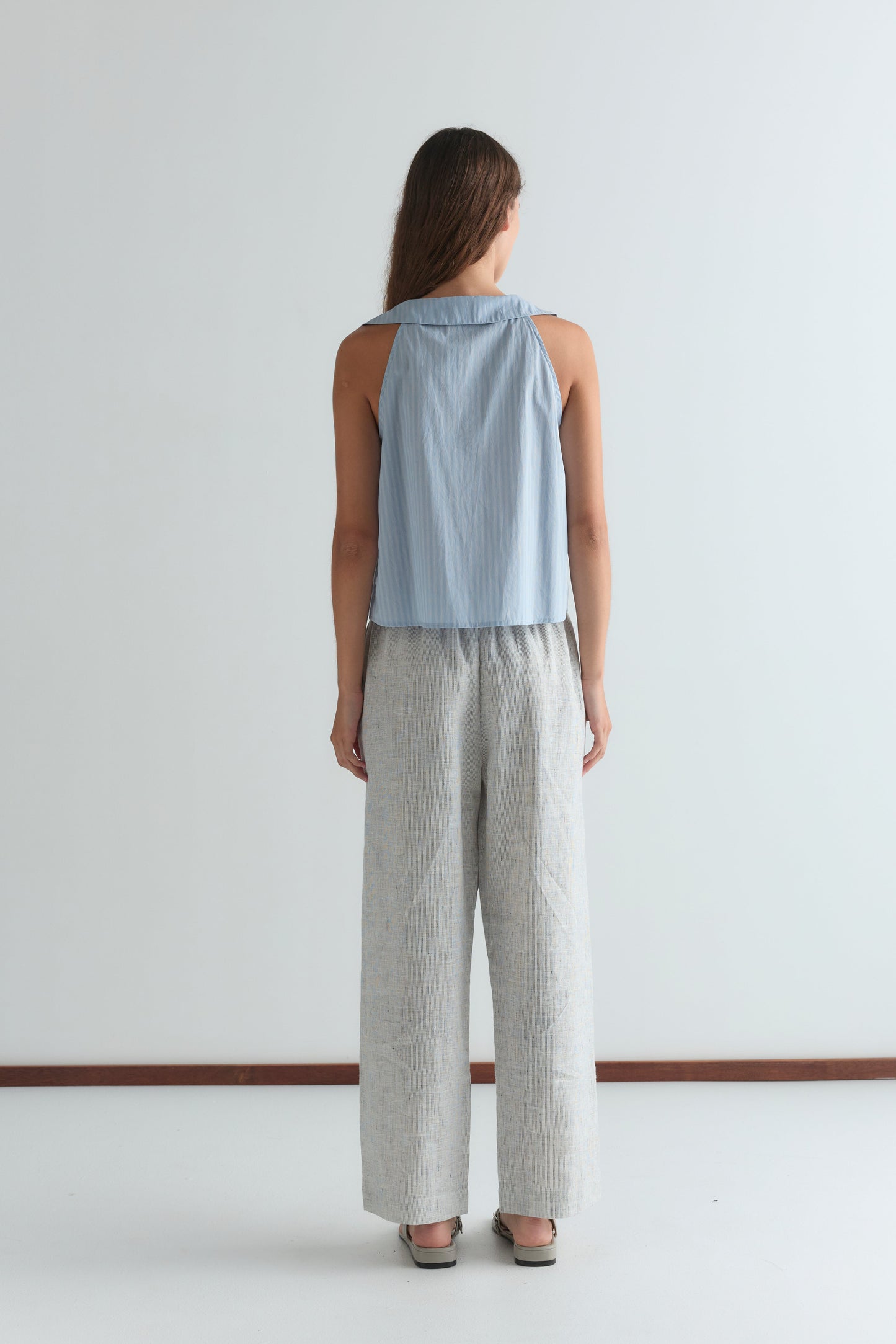A woman faces away from the camera wearing a Deiji Studios Round Collar Top - Lake Blue Stripe, loose light gray patterned pants, and beige sandals, standing against a plain white wall.