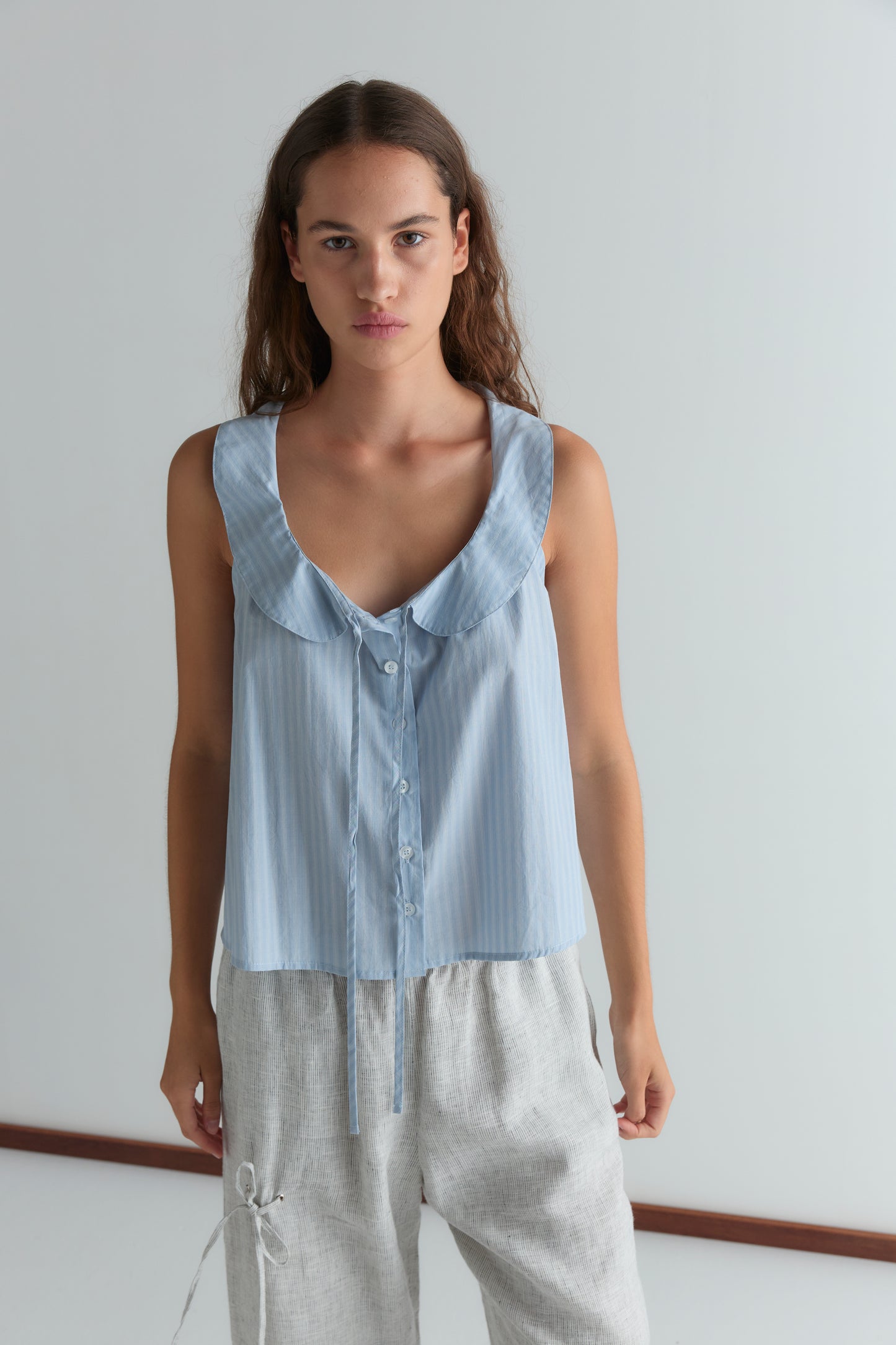 A woman with long, wavy brown hair and a neutral expression stands against a white background wearing a light blue sleeveless blouse with a ruffled collar and buttons, paired with Deiji Studios Eyelet Pant - Fine Stripe.