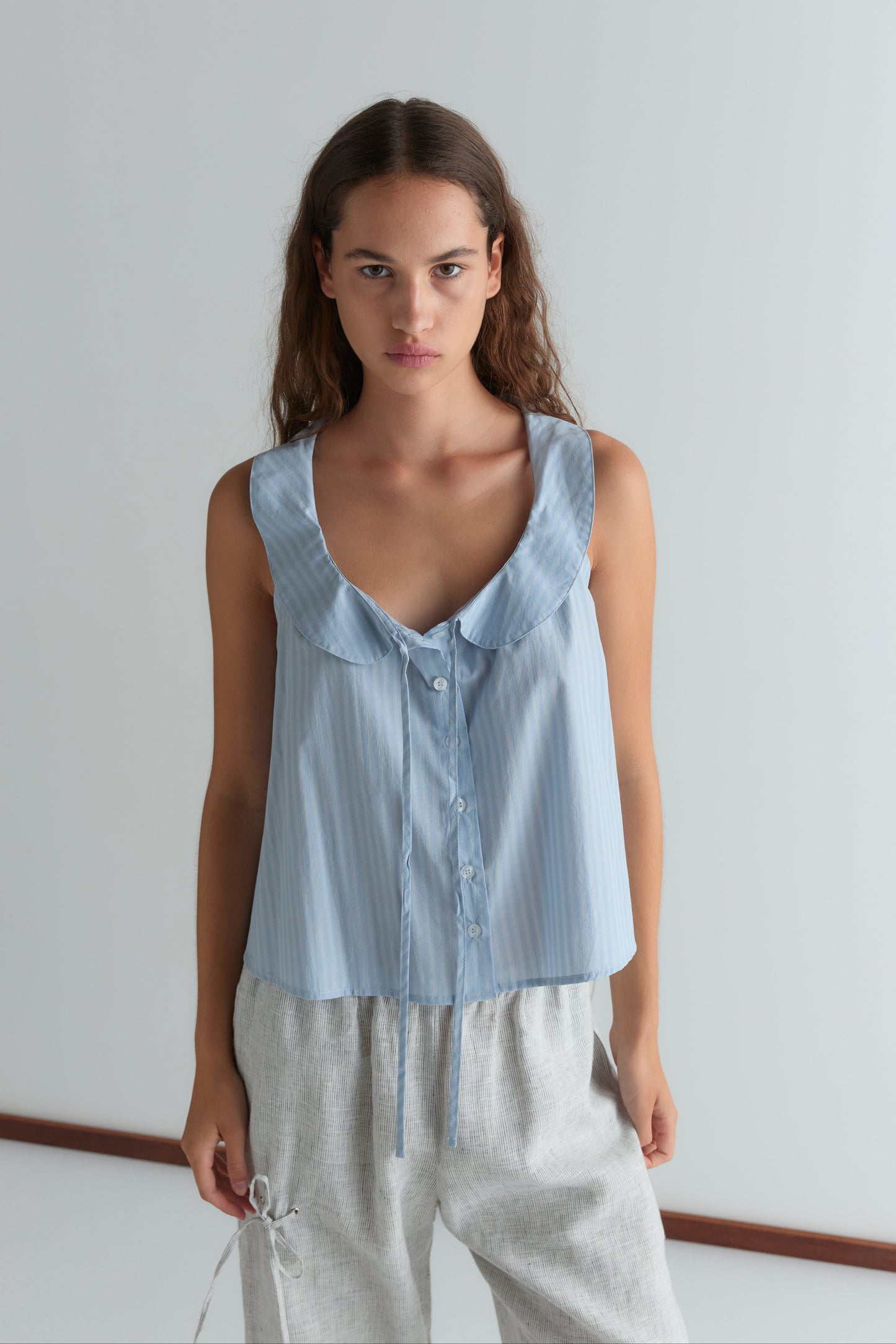 A young woman with long brown hair stands in a bright room, wearing Deiji Studios’ Round Collar Top in Lake Blue Stripe and light gray drawstring pants, looking directly at the camera with a neutral expression.