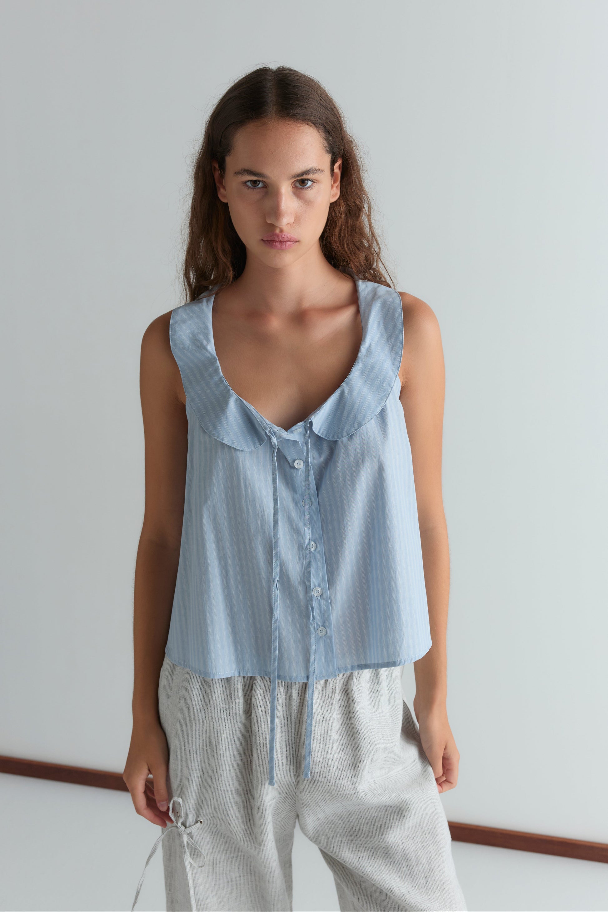 A young woman with long brown hair stands in a bright room, wearing Deiji Studios’ Round Collar Top in Lake Blue Stripe and light gray drawstring pants, looking directly at the camera with a neutral expression.