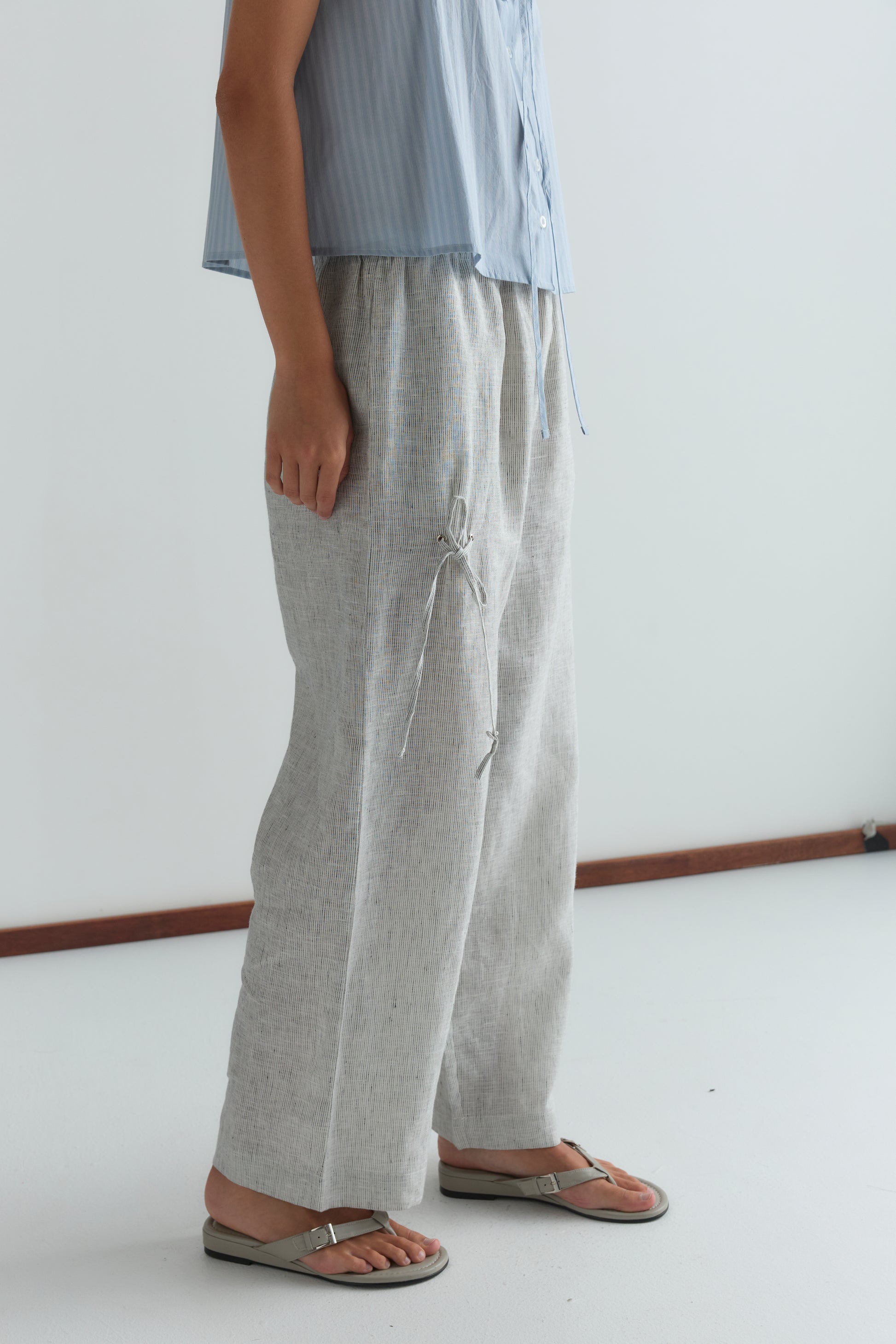 A person stands indoors in a light blue short-sleeved shirt, Deiji Studios Eyelet Pant - Fine Stripe in light gray, and silver flip-flops against a simple, minimalistic background.