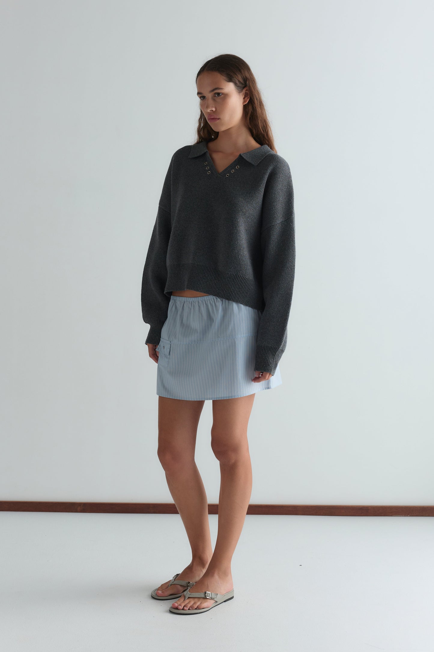 A woman stands indoors wearing a dark gray long-sleeve sweater with a collar, beige sandals, and the Pocket Skirt in Lake Blue Stripe. She has long brown hair, a neutral expression, and poses against a plain white background.