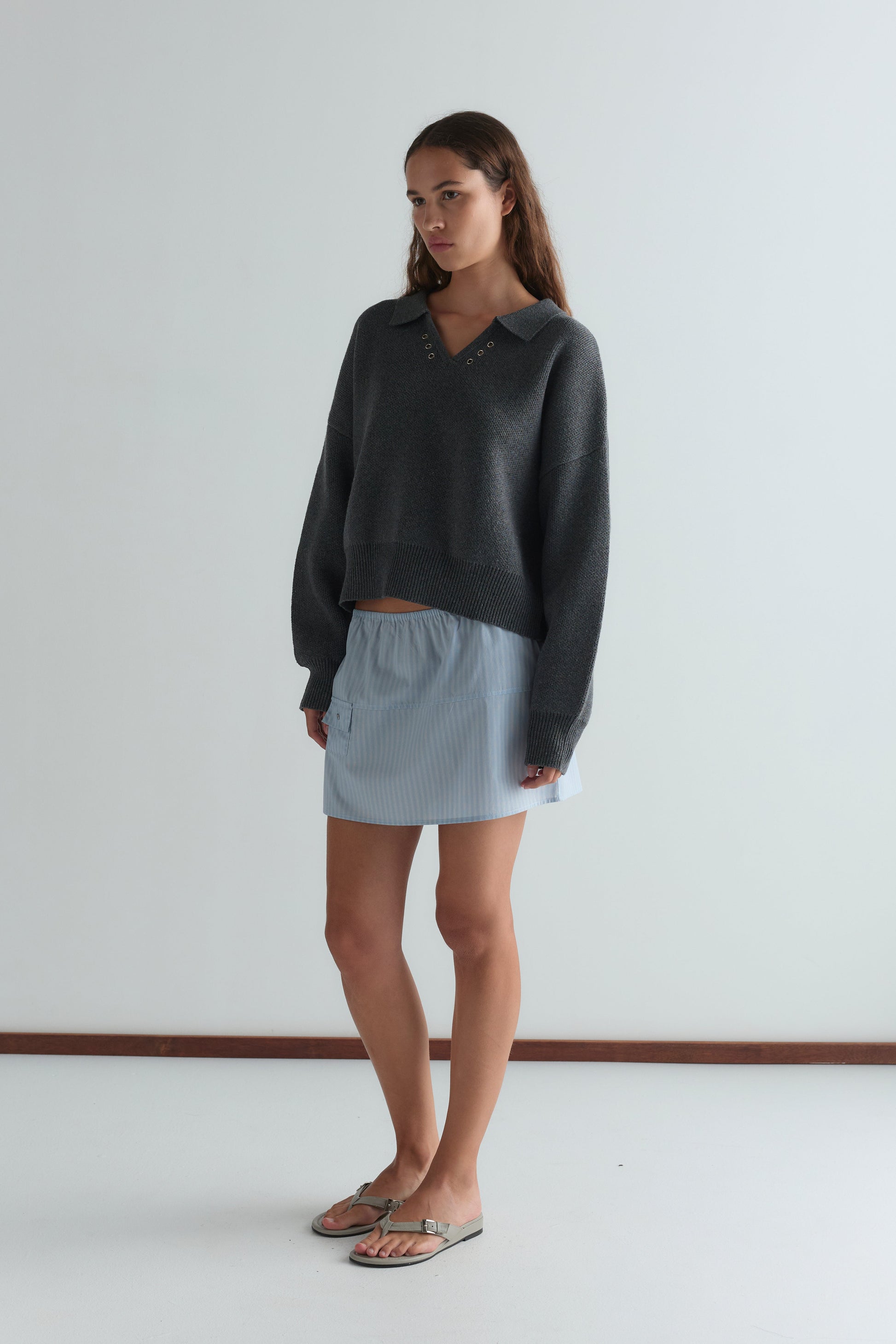 A woman stands indoors wearing a dark gray long-sleeve sweater with a collar, beige sandals, and the Pocket Skirt in Lake Blue Stripe. She has long brown hair, a neutral expression, and poses against a plain white background.