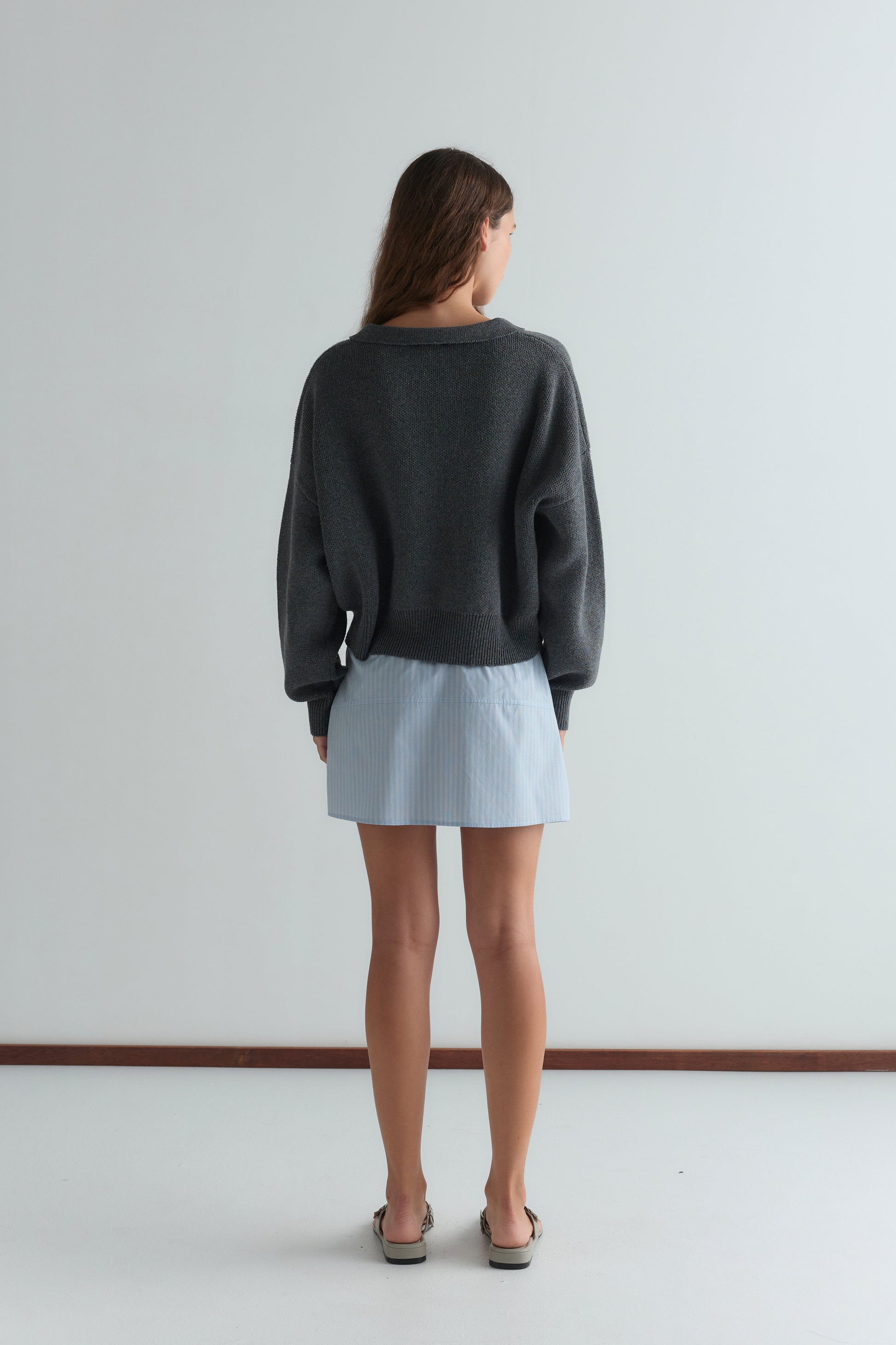 A woman faces away from the camera, wearing the Eyelet Knit - Dark Pebble by Deiji Studios, a light blue pinstriped mini skirt, and sandals in a minimal, light-toned room with a white floor and wall.