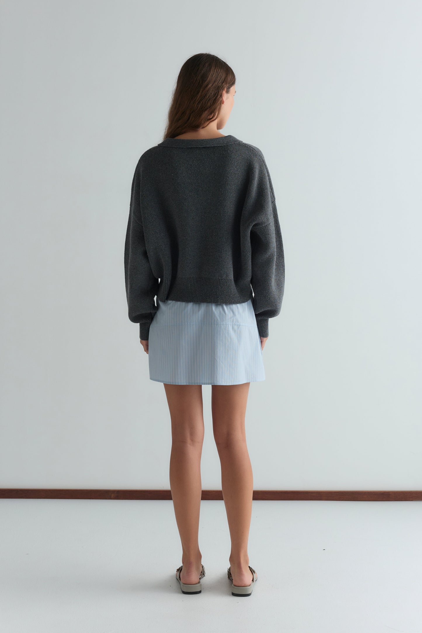 A woman faces away, wearing a loose gray sweater, the Pocket Skirt - Lake Blue Stripe, and sandals, standing against a plain white wall.