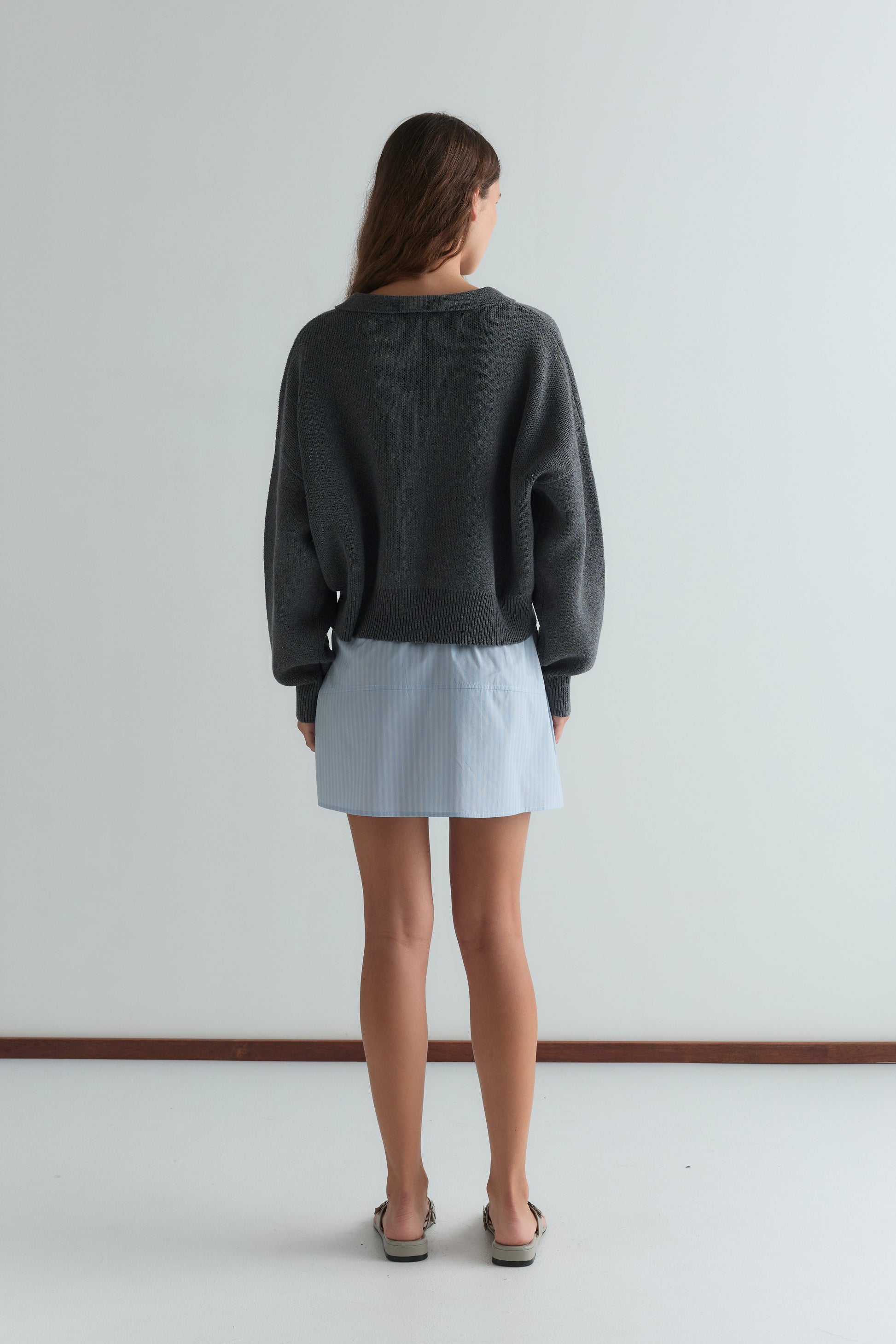 A woman faces away, wearing a loose gray sweater, the Pocket Skirt - Lake Blue Stripe, and sandals, standing against a plain white wall.