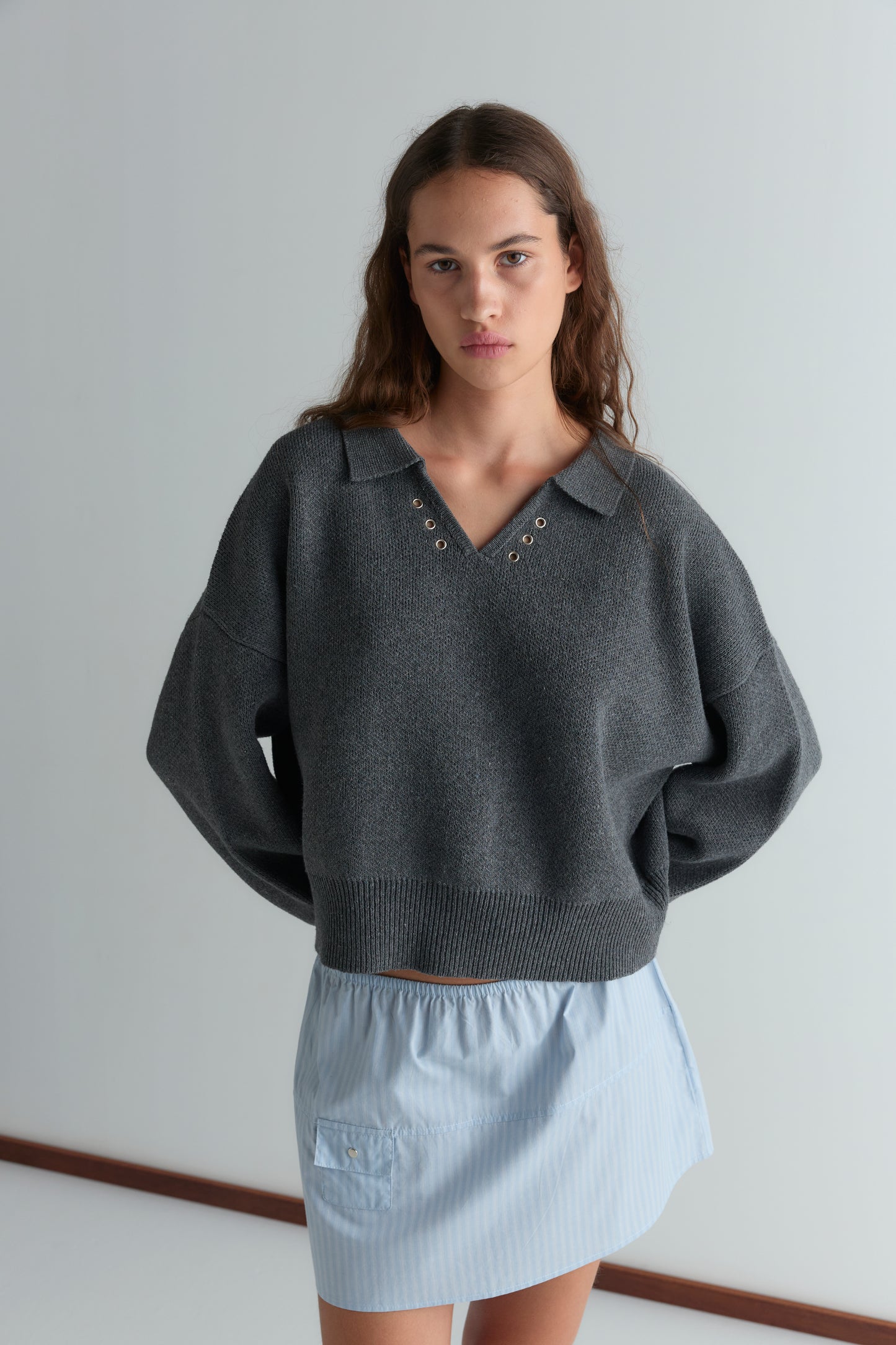 A young woman with long brown hair wears the Eyelet Knit - Dark Pebble sweater by Deiji Studios with a light blue mini skirt, standing against a plain background and looking at the camera.