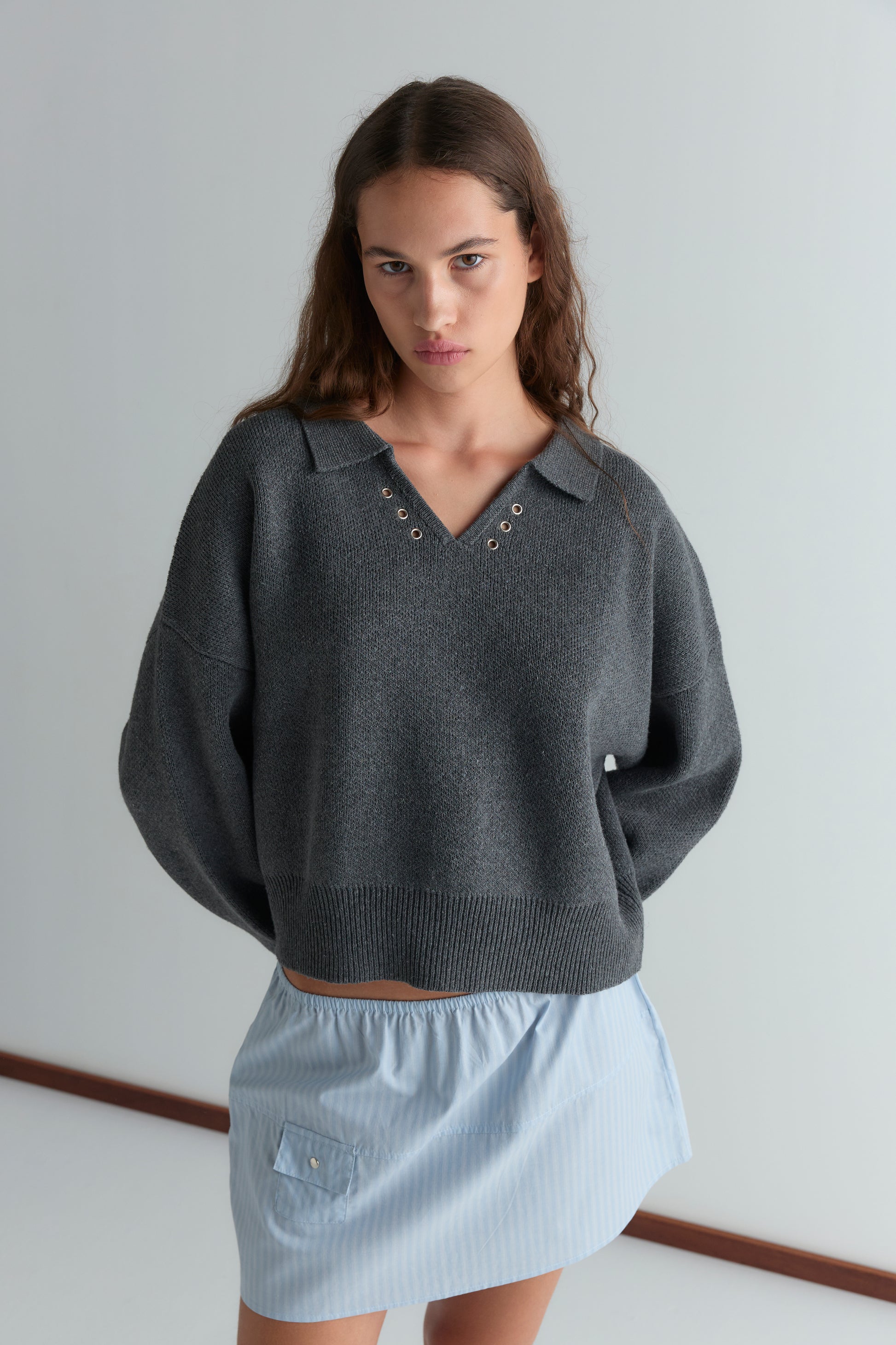 A woman with long brown hair wears a loose dark gray V-neck sweater with grommet details and the Pocket Skirt - Lake Blue Stripe, standing against a plain white wall.