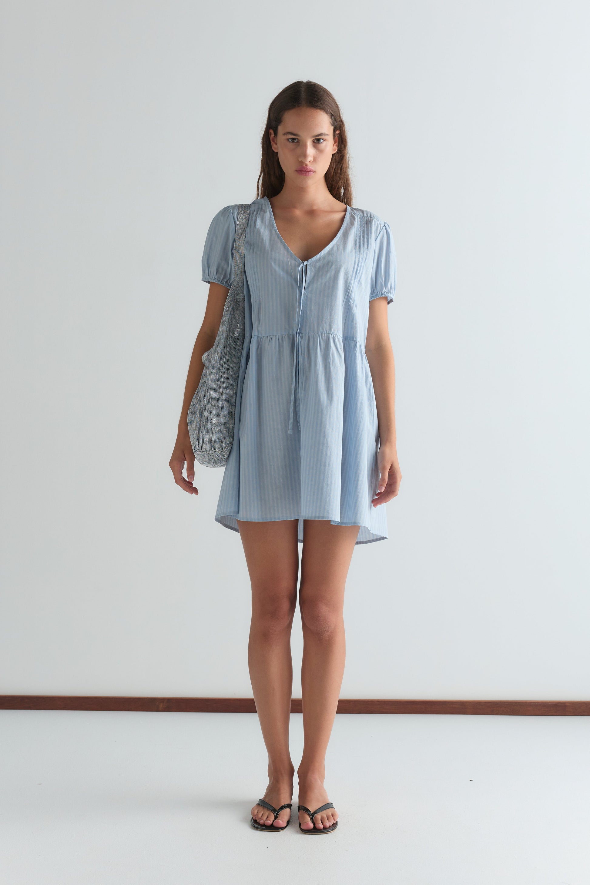 A woman stands facing forward against a plain white background, wearing the V Pleat Dress - Lake Blue Stripe by Deiji Studios with short sleeves and knee-length hem. She carries a textured shoulder bag and wears black sandals.