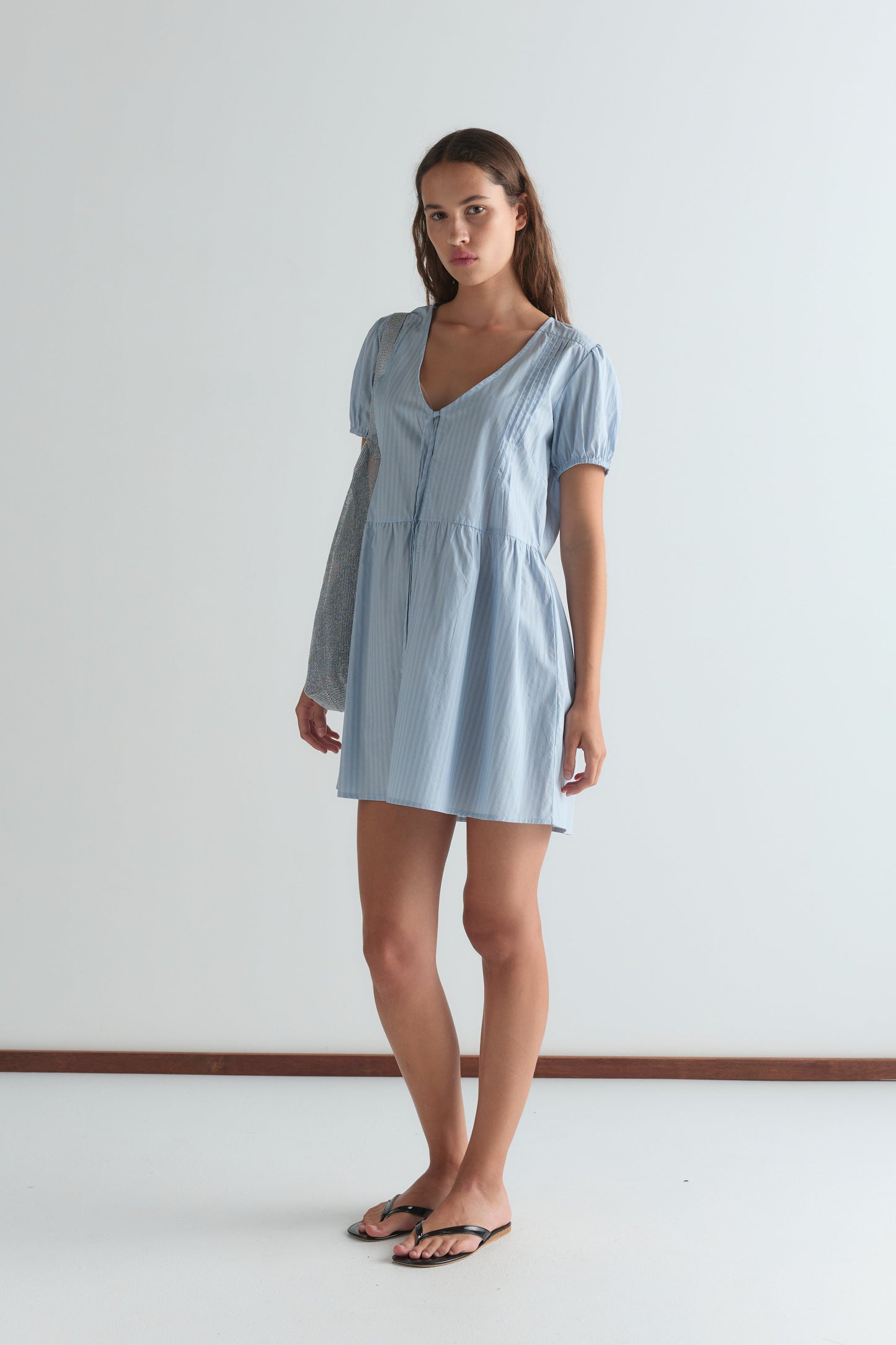 A woman with long brown hair and a neutral expression stands against a white wall, wearing the V Pleat Dress - Lake Blue Stripe from Deiji Studios with short sleeves, a v-neckline, and flip flops.