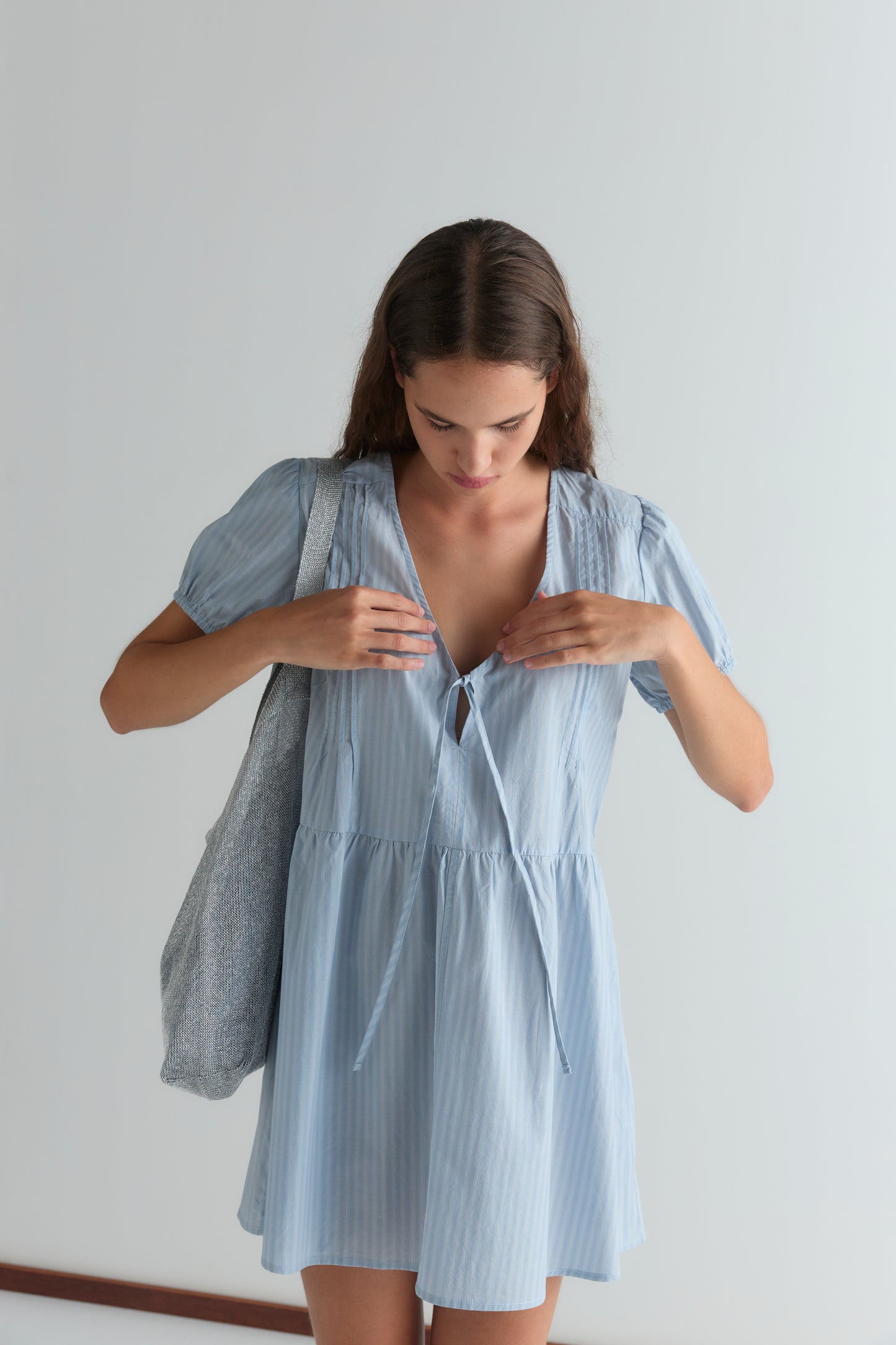 A woman with long hair stands against a plain background, looking down as she adjusts the front tie of her light blue, short-sleeved V Pleat Dress. She carries a textured gray tote bag on her shoulder.
